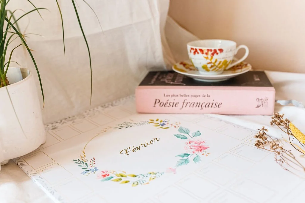 Nature morte chaleureuse avec calendrier de février fleuri, livres de poésie française, tasse de thé vintage et plante en pot.