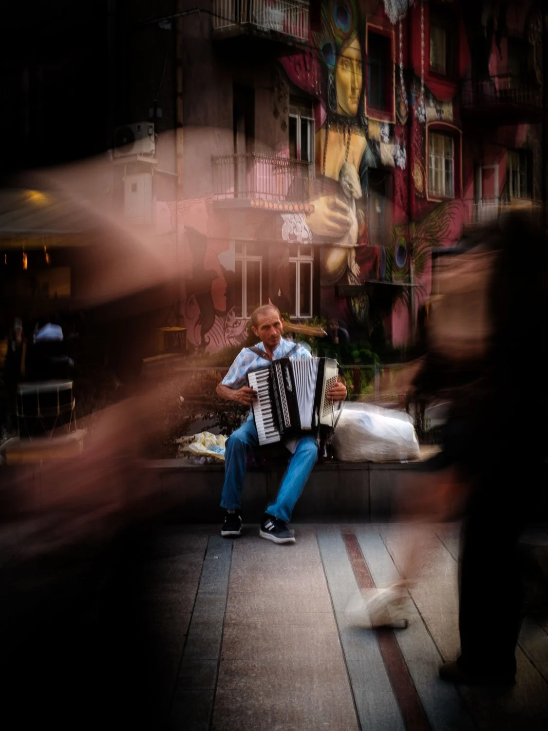 Street musician playing an accordion in front of colorful mural-covered buildings, with blurred pedestrians passing by