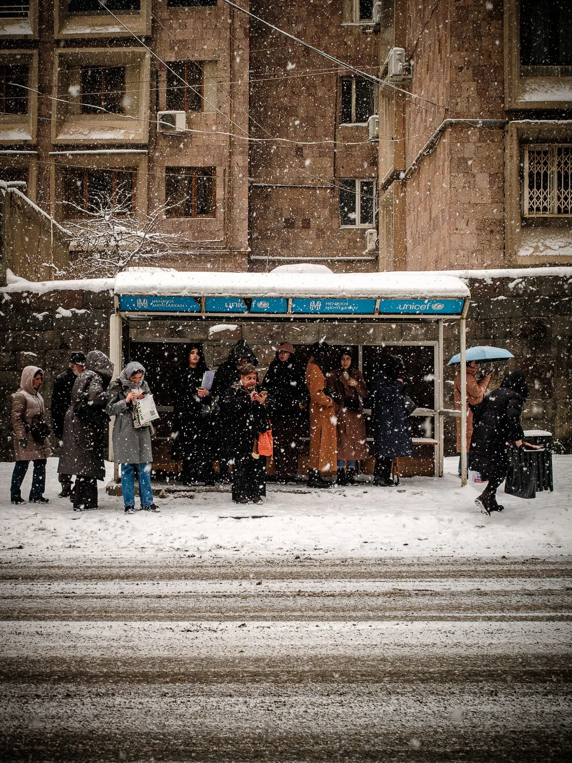 People shelter under a UNICEF bus stop during a heavy snowfall in an urban neighborhood.