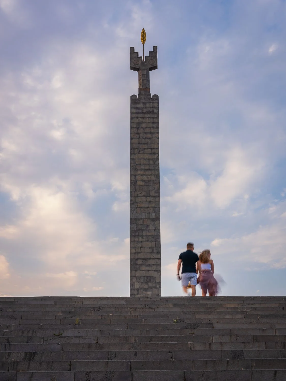 Stone monument topped with a golden leaf emblem, viewed above broad steps with a couple walking beneath a cloudy evening sky