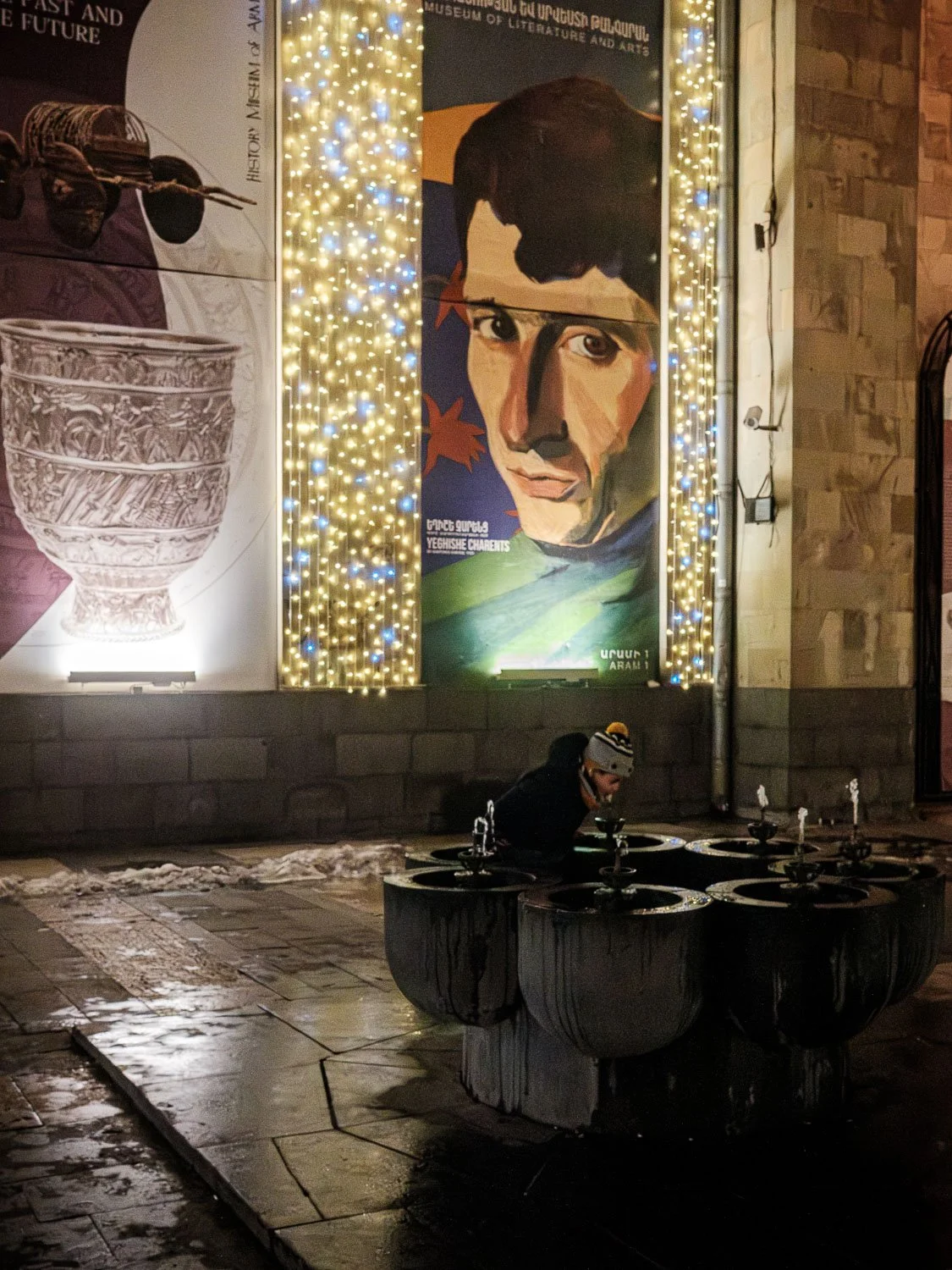 Child drinking from a public fountain outside a brightly lit museum with colorful historical posters.