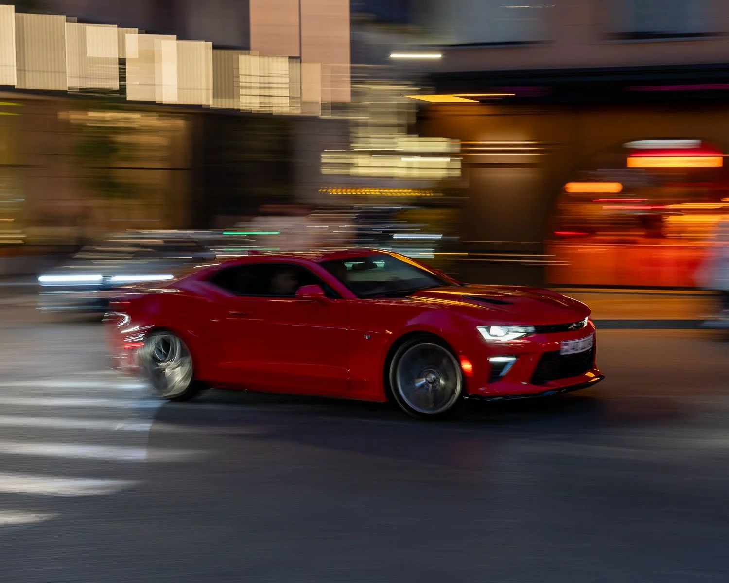 Red Chevrolet Camaro driving through a city at night with motion-blurred lights and traffic in the background