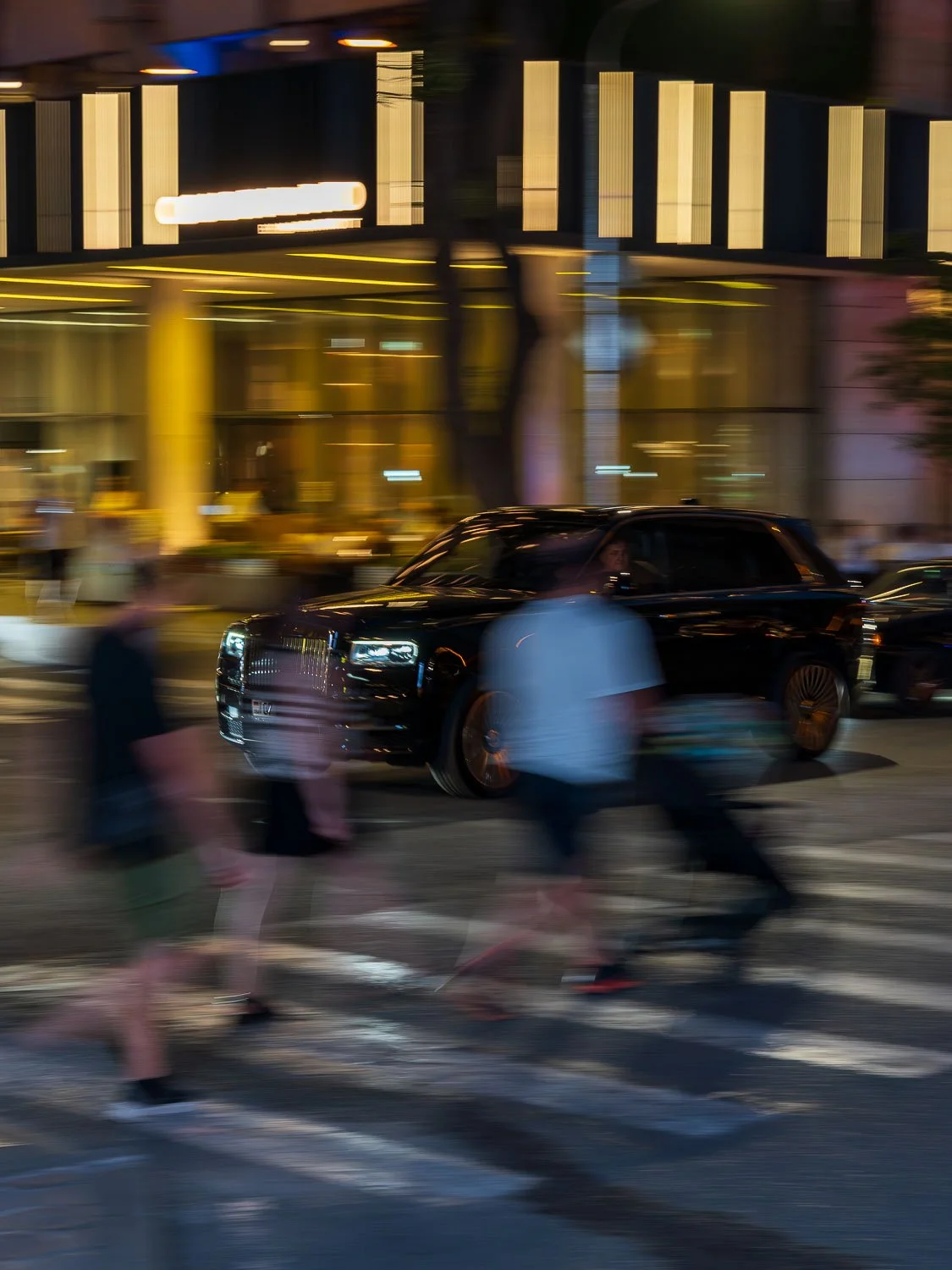 Motion-blurred pedestrians crossing in front of a black luxury SUV at night outside a brightly lit modern building
