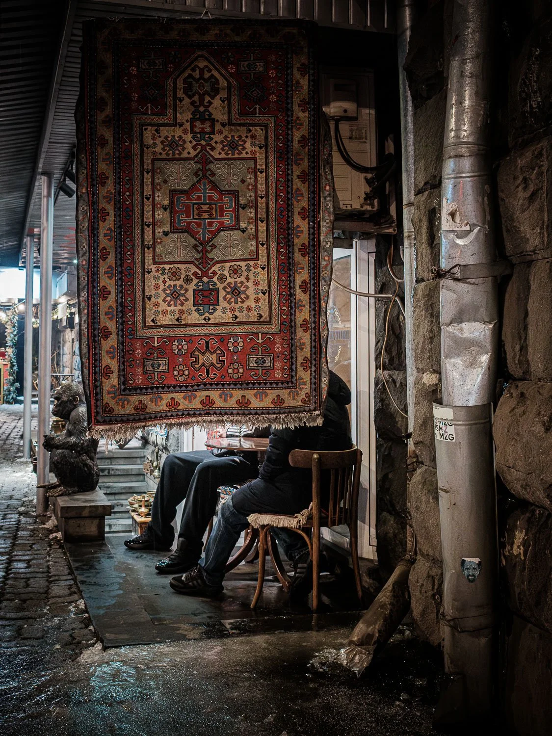 Ornate patterned rug hanging in a dim, narrow market alley beside seated people and exposed plumbing