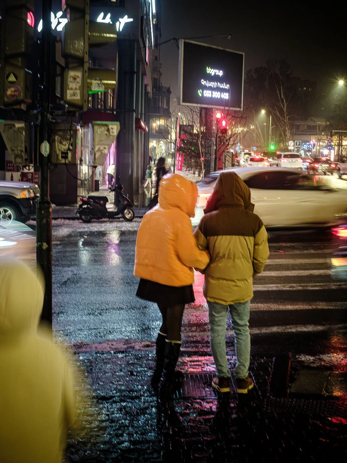 Two people in colorful hooded jackets cross a wet, busy city street at night under illuminated signs and traffic lights.