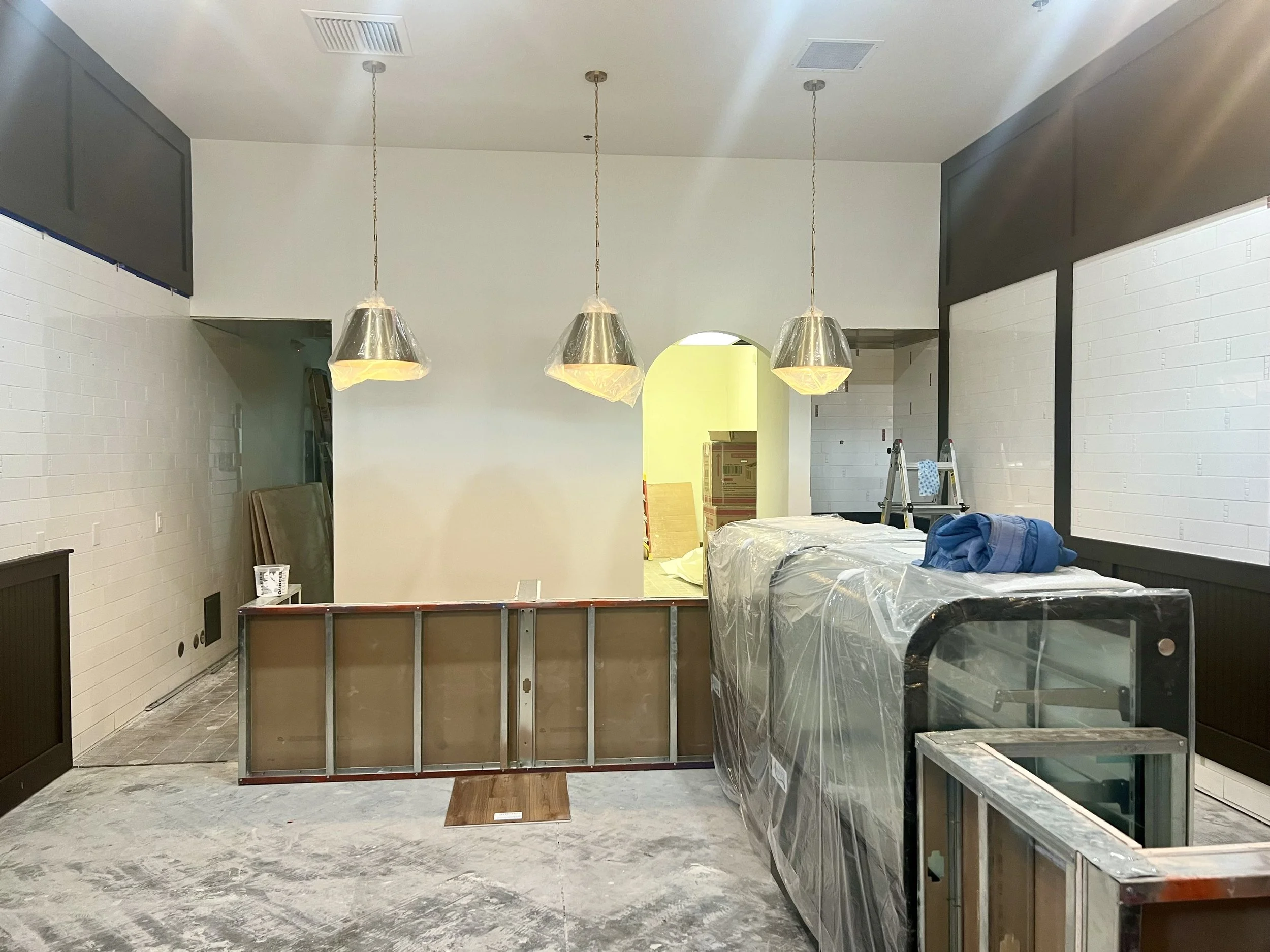 Unfinished restaurant interior under construction with three pendant lights, exposed framing, partially installed counters, and construction materials.