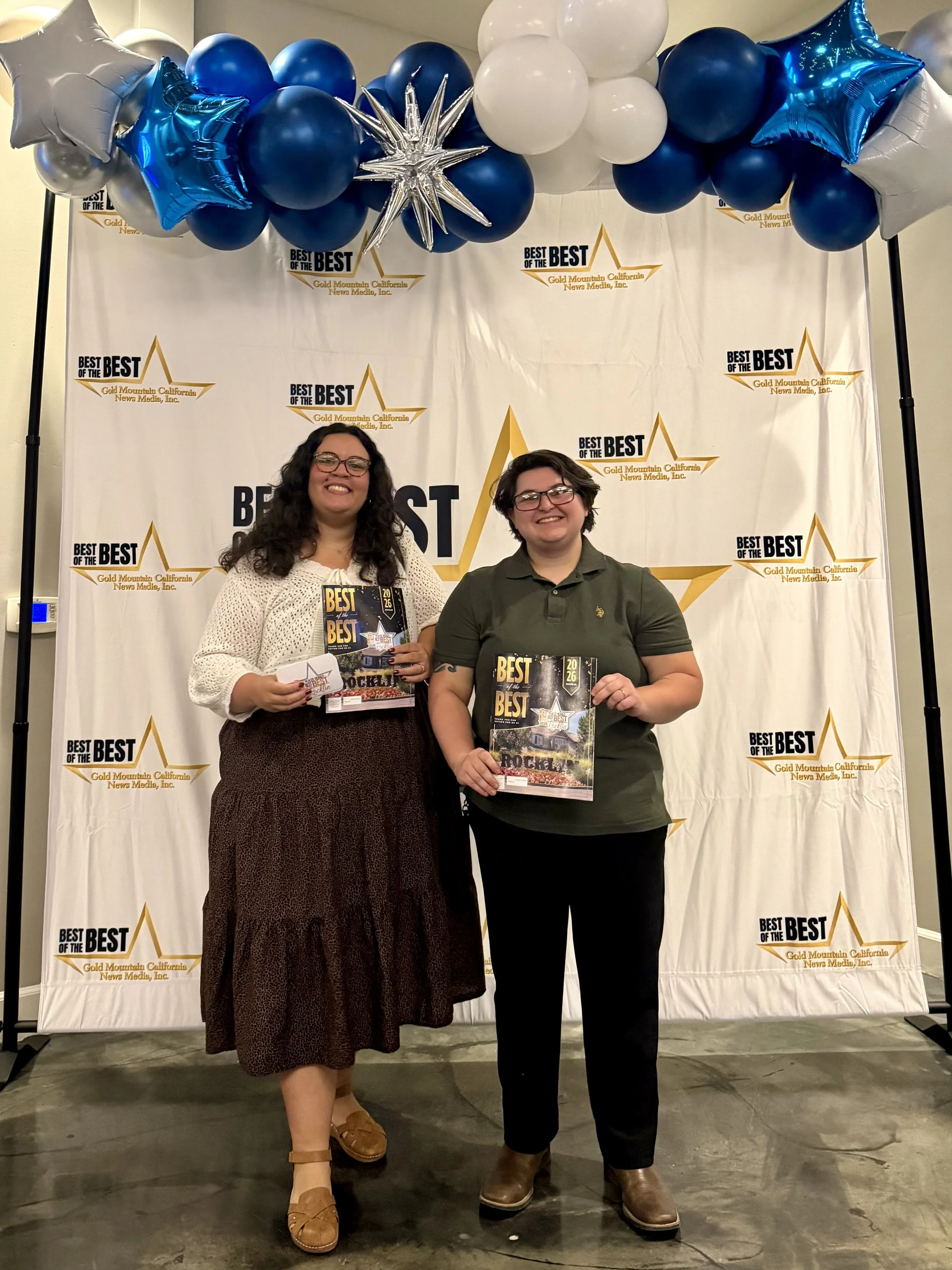 Two women hold Best of the Best awards in front of a blue-and-white balloon display and Gold Mountain California News Media backdrop.