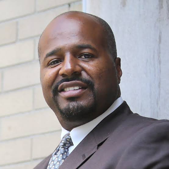 Portrait of a smiling Black man in a dark suit and patterned tie standing against a light brick wall