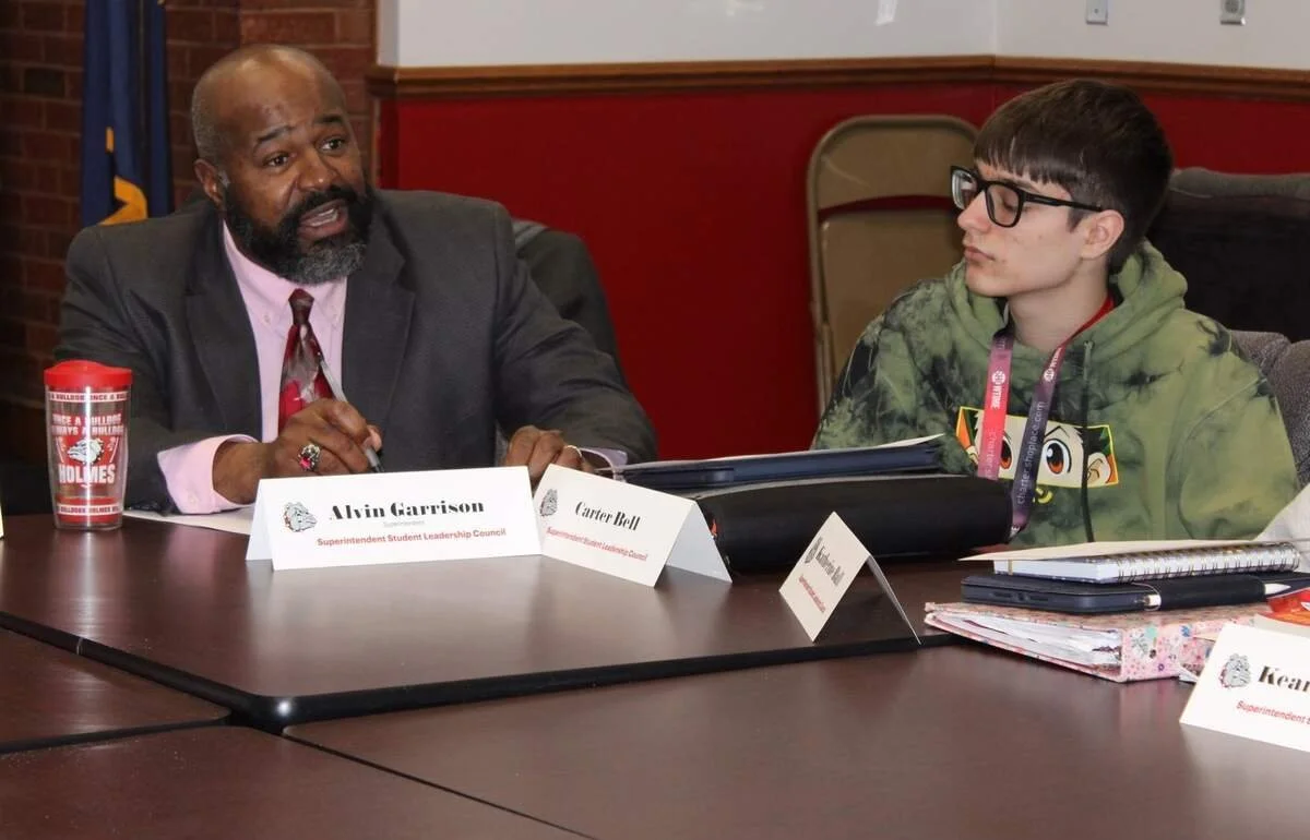 Man in a suit speaking with a student wearing glasses at a formal meeting table, with name placards and notebooks.