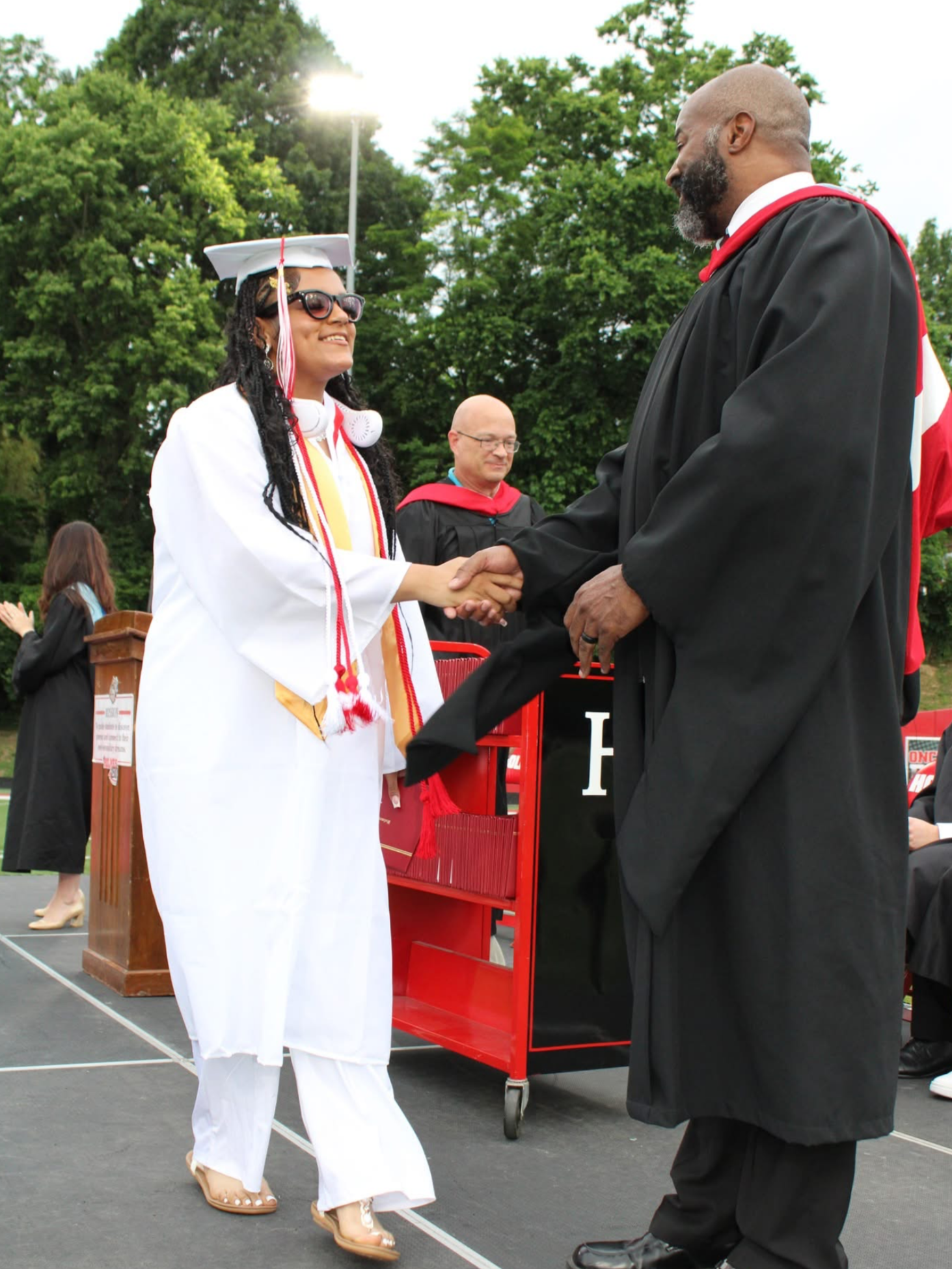 Graduate in white cap and gown shakes hands with an academic official during an outdoor graduation ceremony.