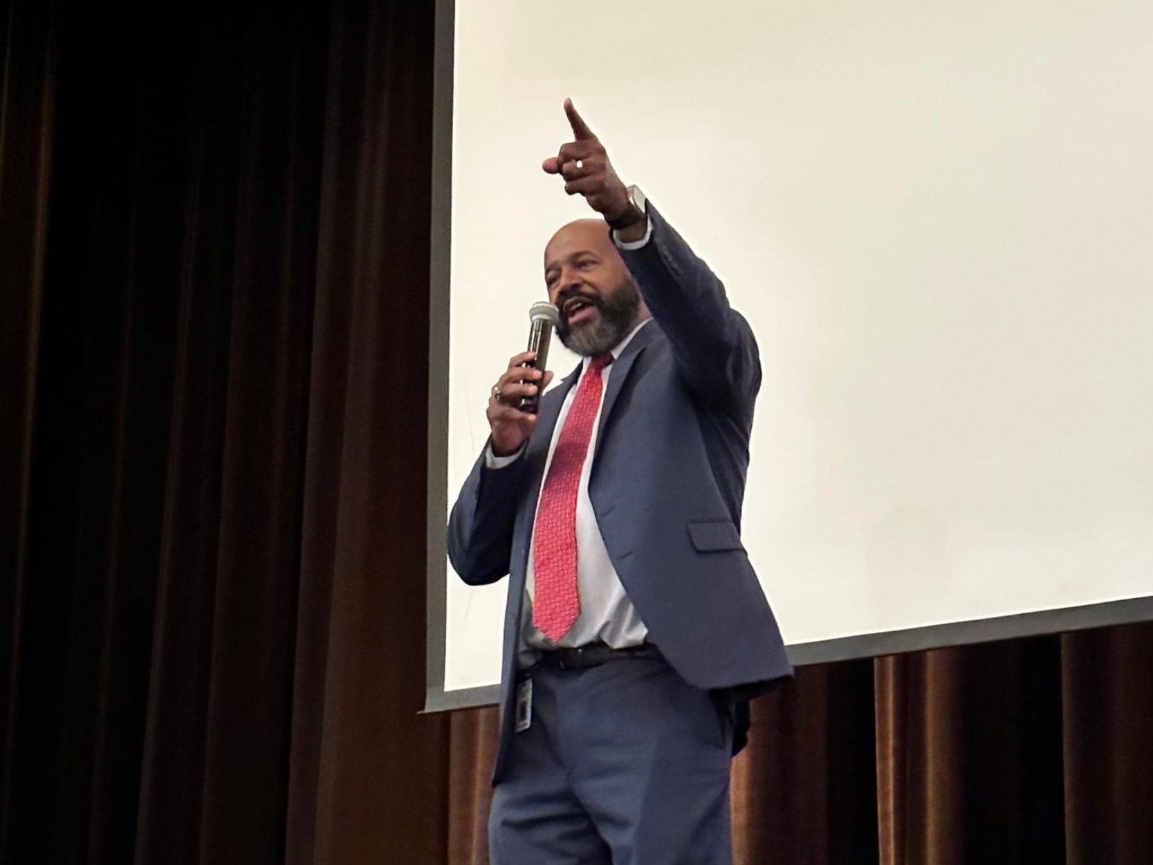 Man in a suit speaking into a microphone and pointing upward on a stage