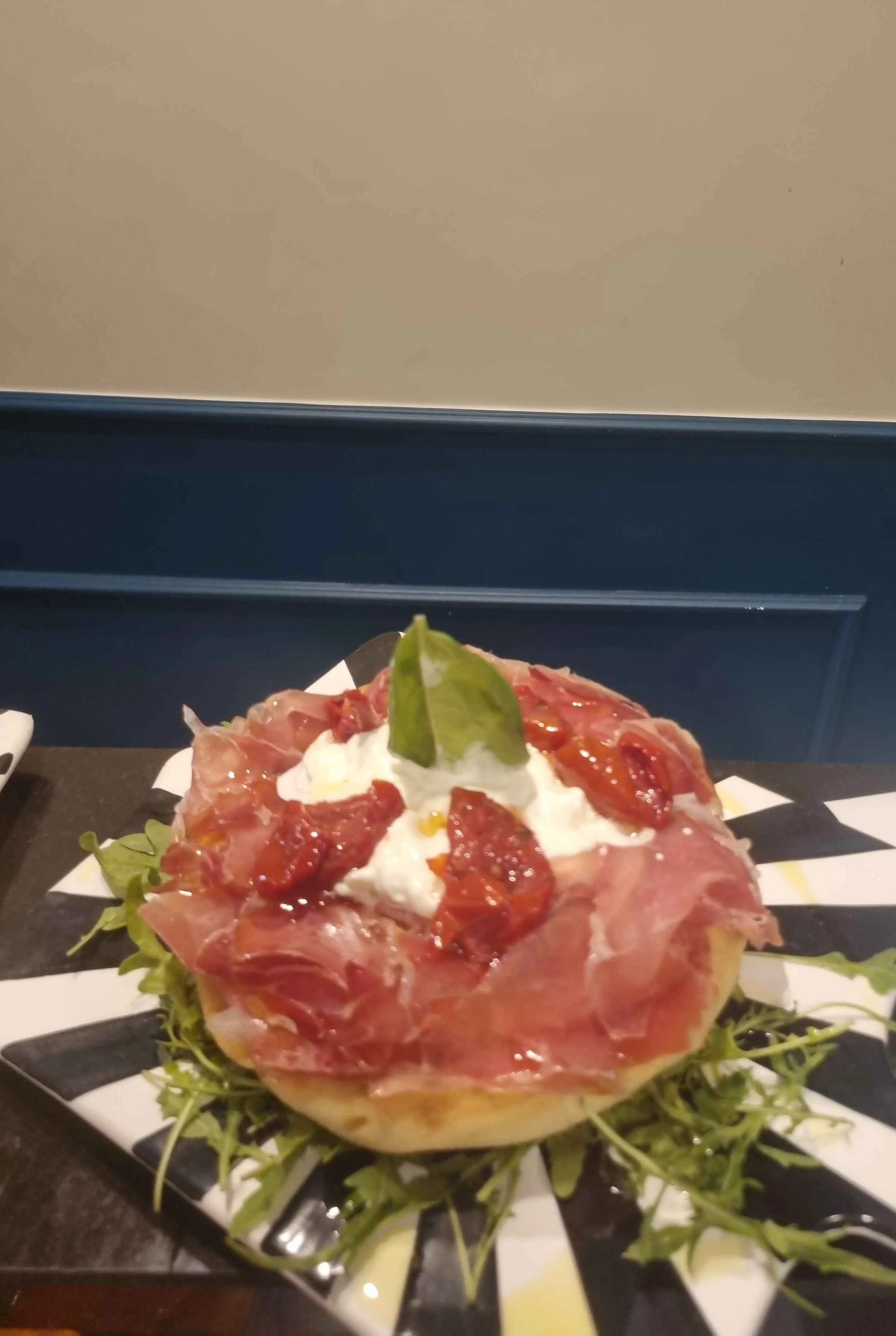 Panino gourmet con prosciutto crudo, pomodori, burrata, rucola e basilico su piatto bianco e nero.