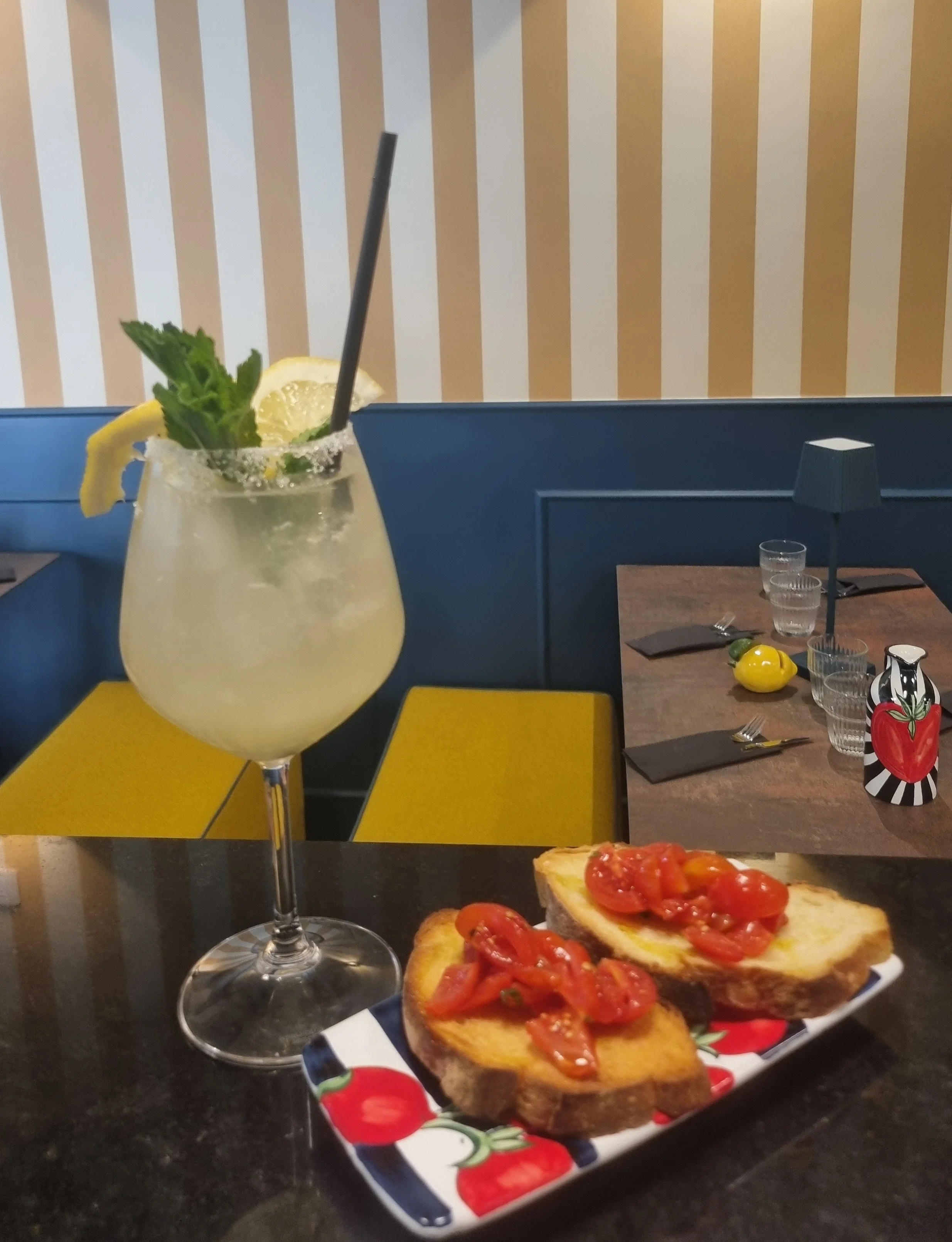 Cocktail al limone con menta e cannuccia nera accanto a crostini con pomodori freschi su un tavolo da ristorante.