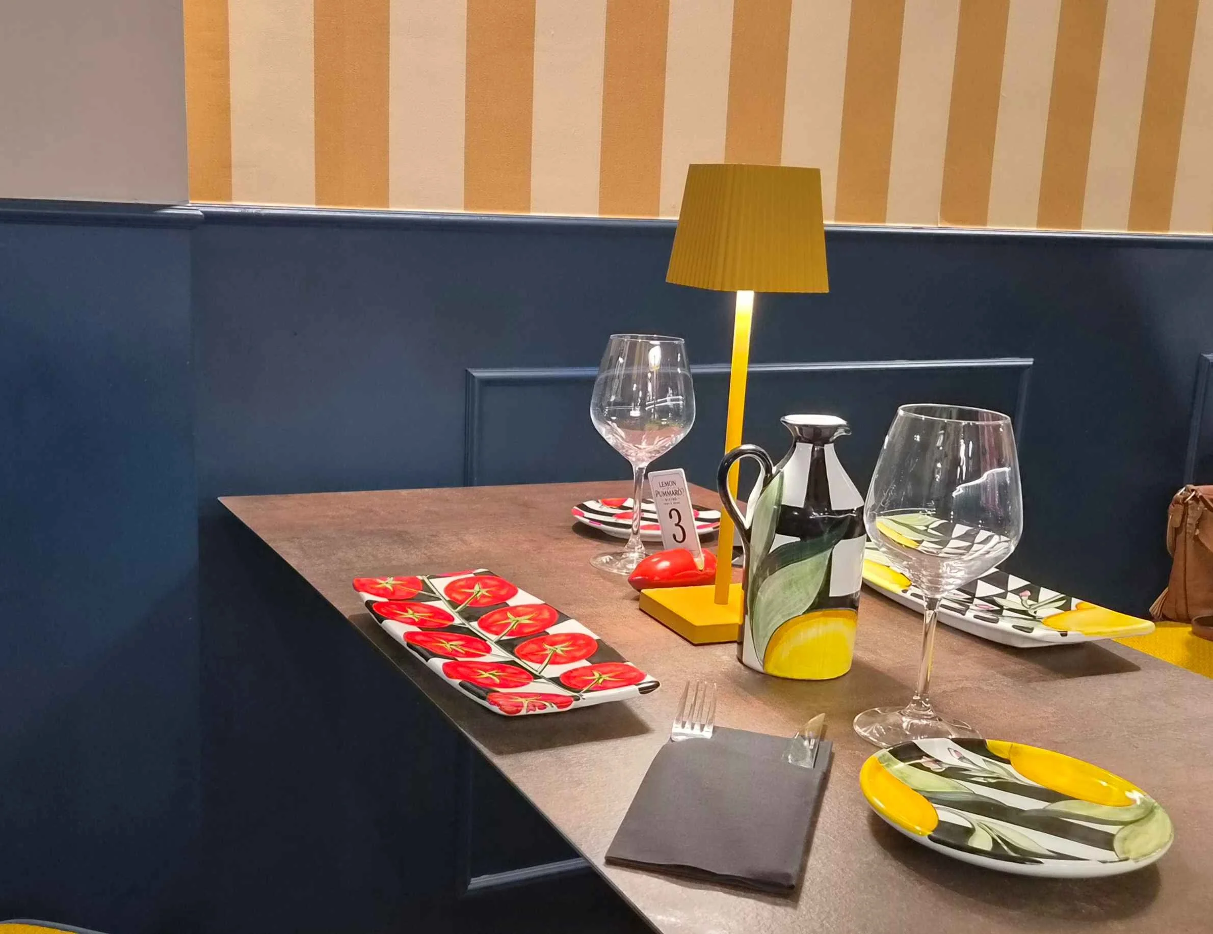 Tavola apparecchiata in un ristorante con piatti colorati, bicchieri, caraffa decorata e lampada gialla.