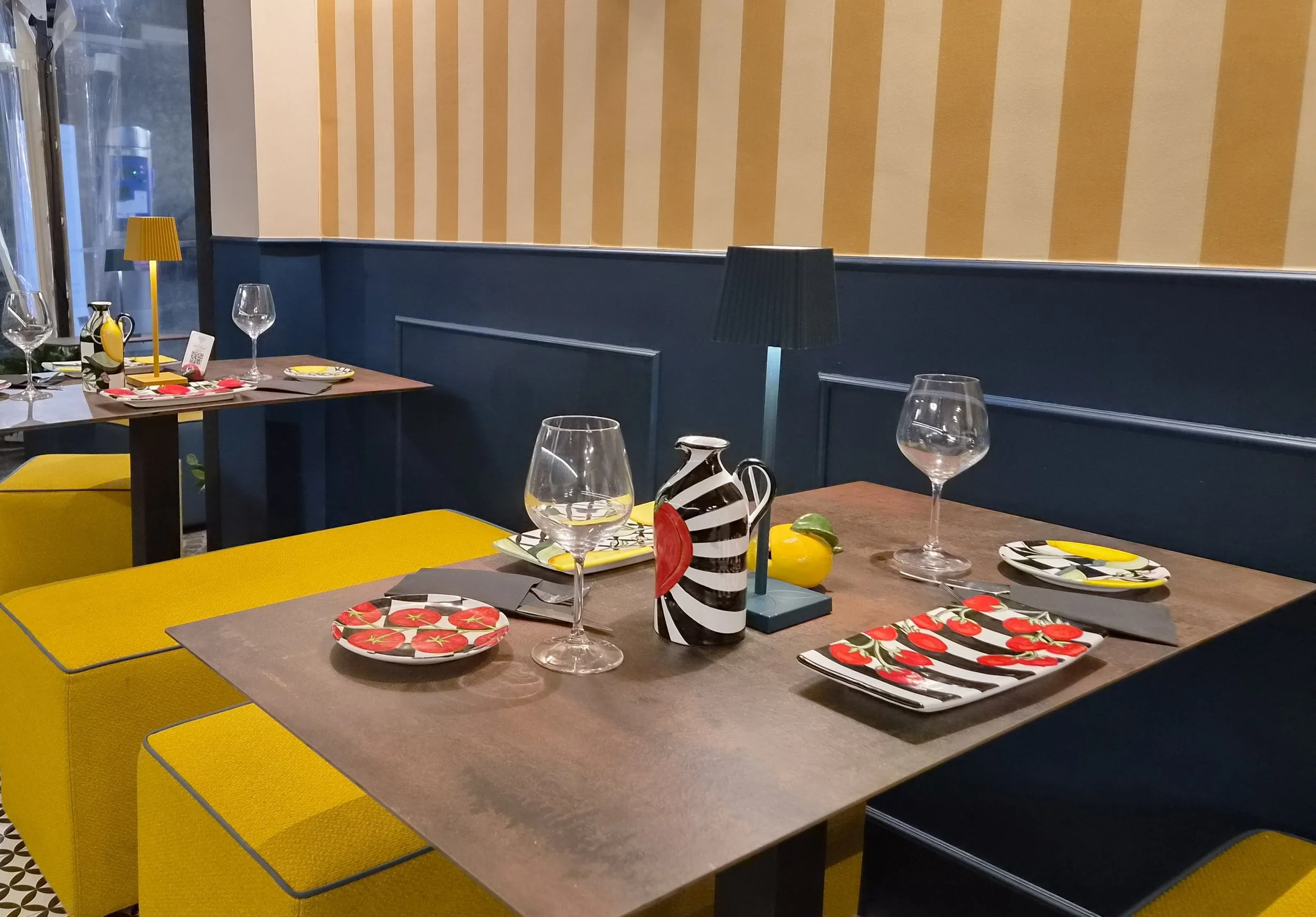 Sala bistro elegante con tavoli apparecchiati, calici da vino, piatti decorati con pomodori e arredi gialli e blu.