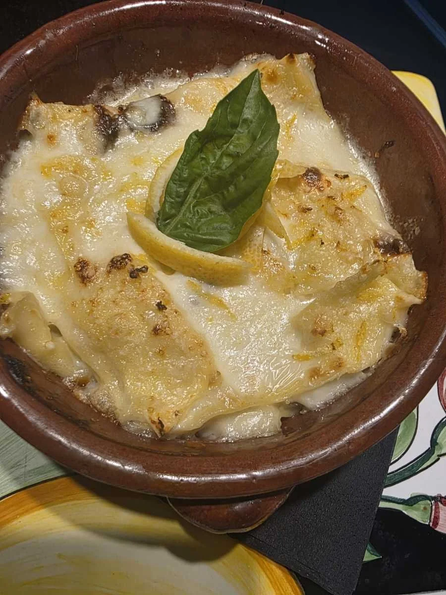 Lasagne al forno cremose con formaggio gratinato, guarnite con foglia di basilico e scorza di limone in una pirofila di terracotta.