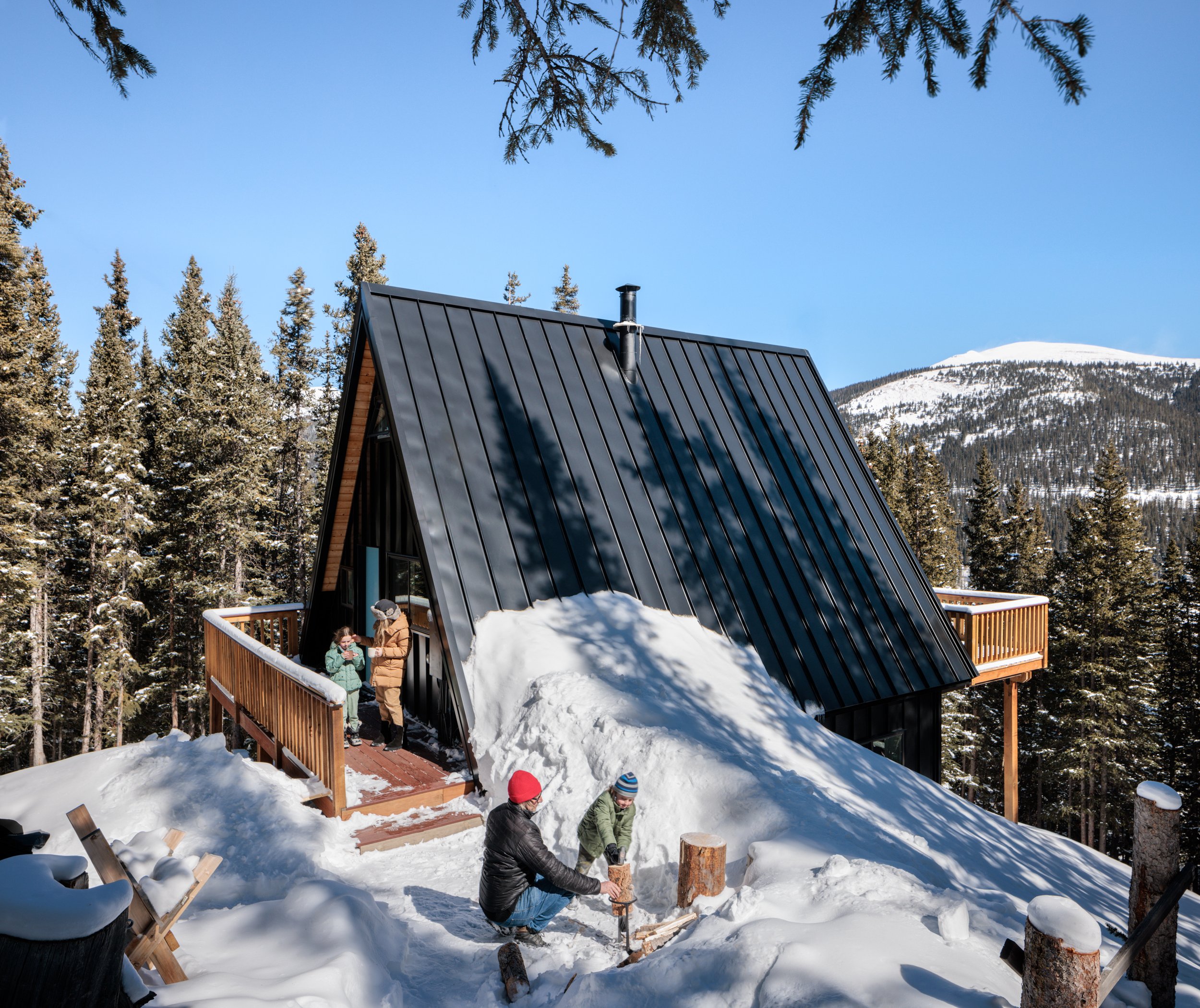 Snow-covered A-frame cabin in a pine forest, with people outside.