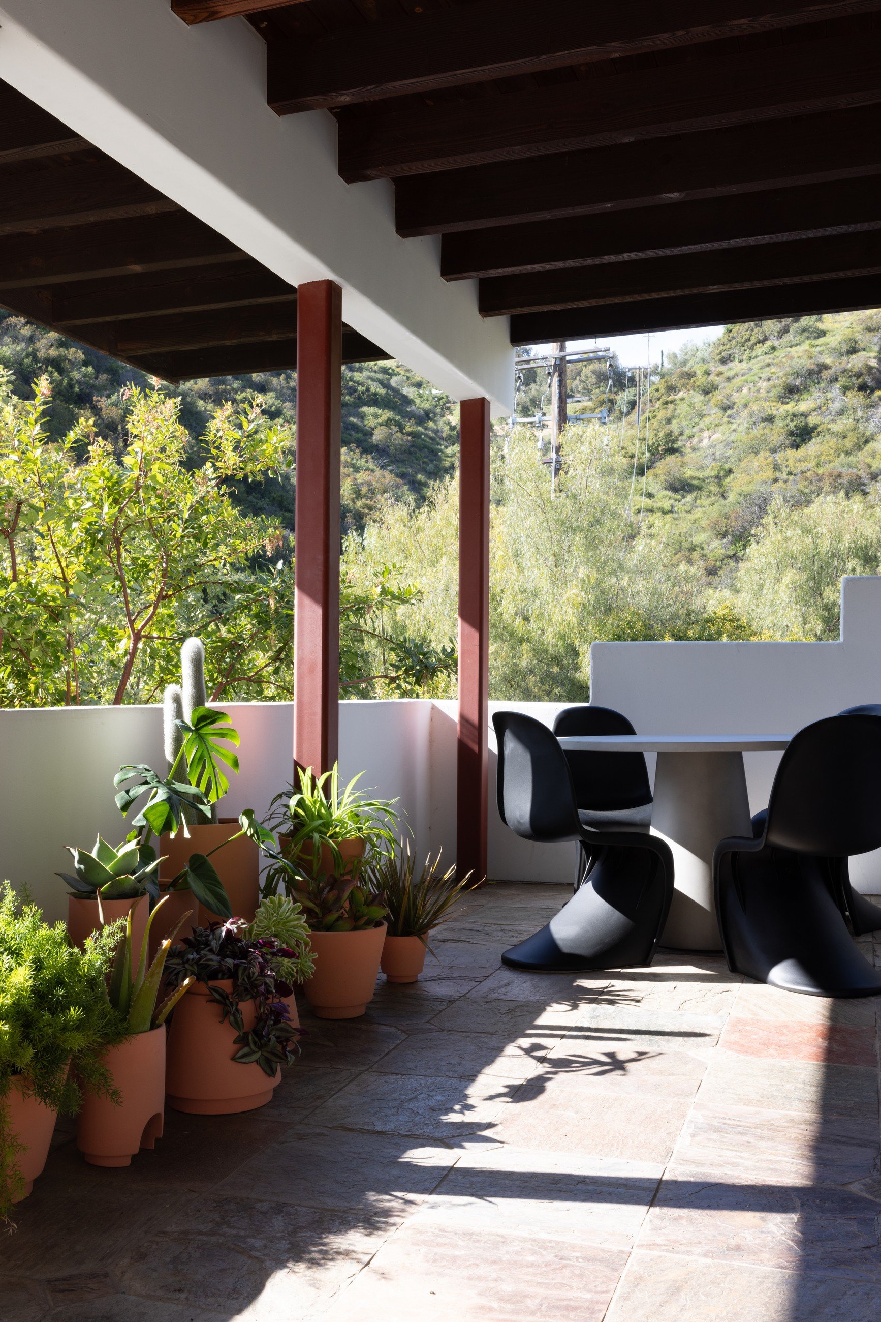 Terracotta potted plants and black sculptural seating on a shaded porch beneath dark timber beams.