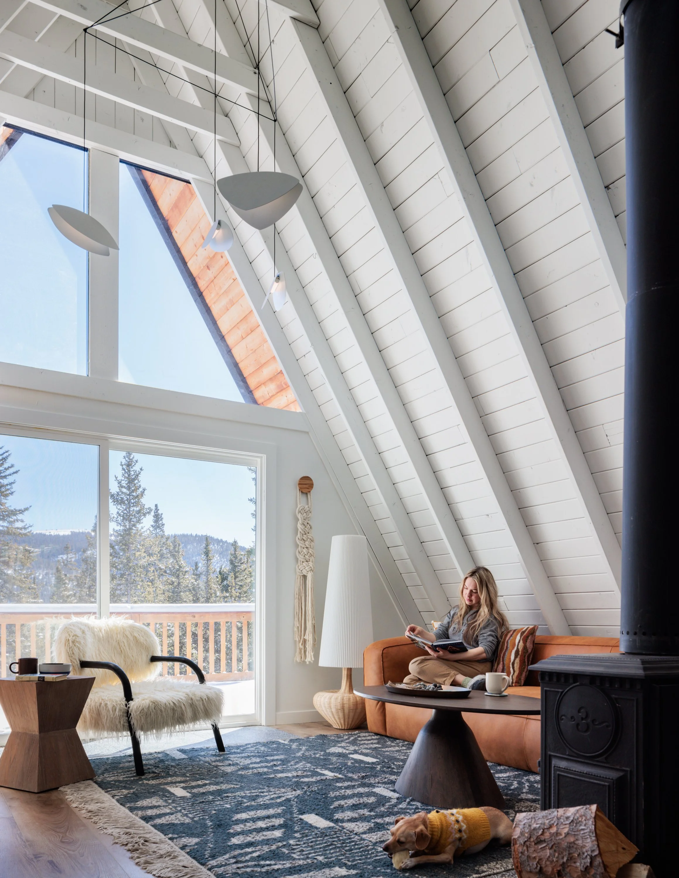 A-frame living room with a vaulted white ceiling, mountain views, and a woman seated beside a dog.