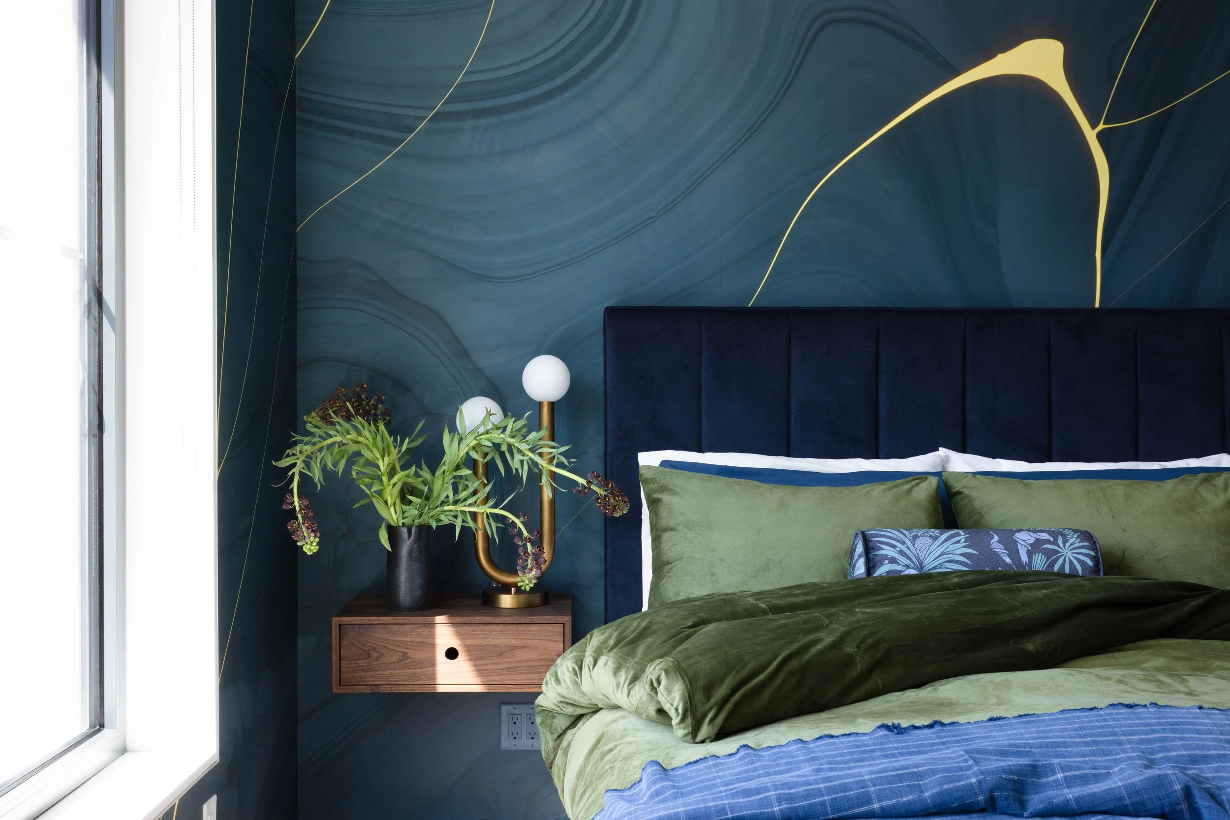 Bedroom with a dark blue upholstered headboard, green bedding and a sculptural wall mural.