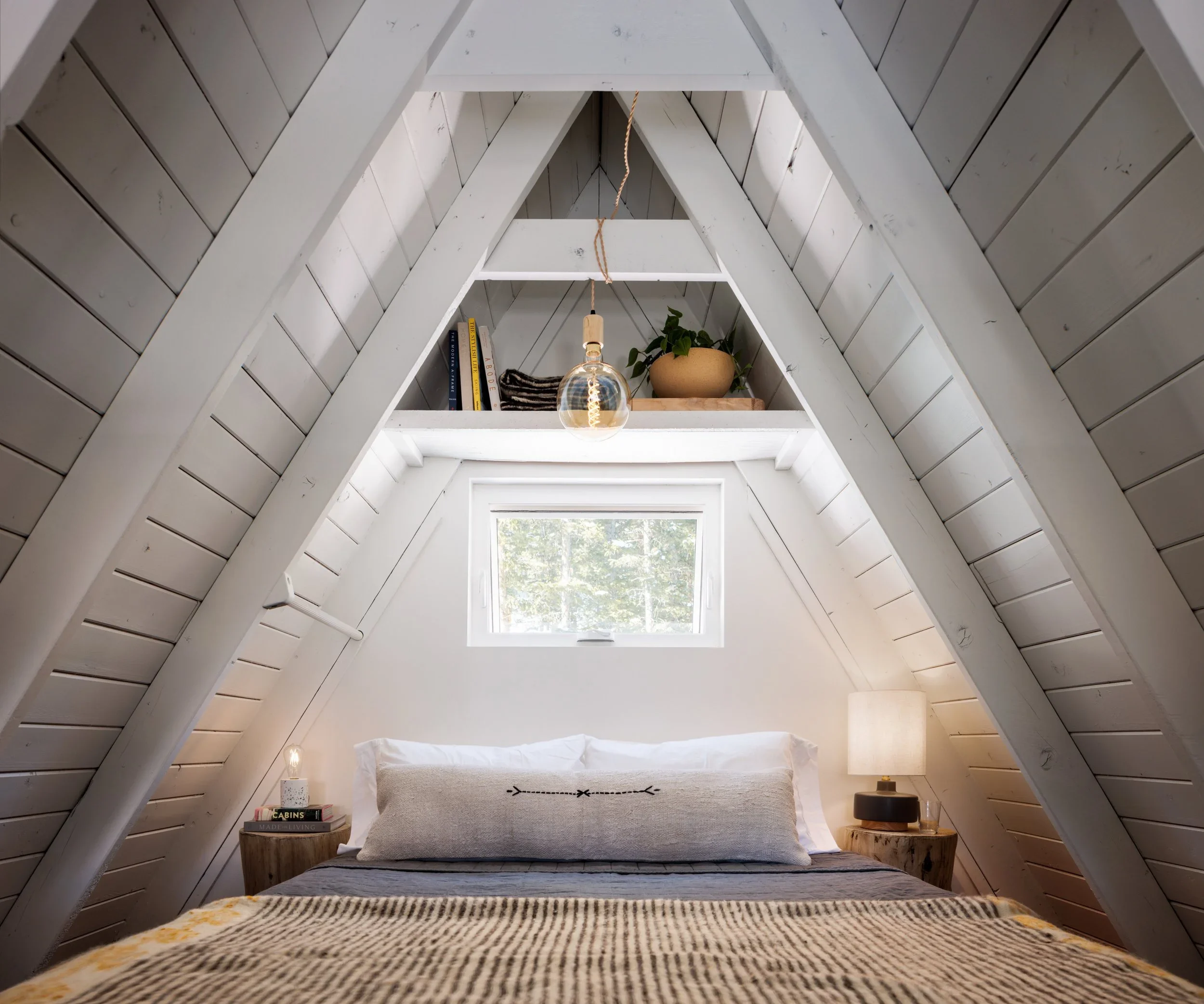 Loft bedroom beneath white A-frame beams with layered neutral bedding, bedside lamps, and a small window.