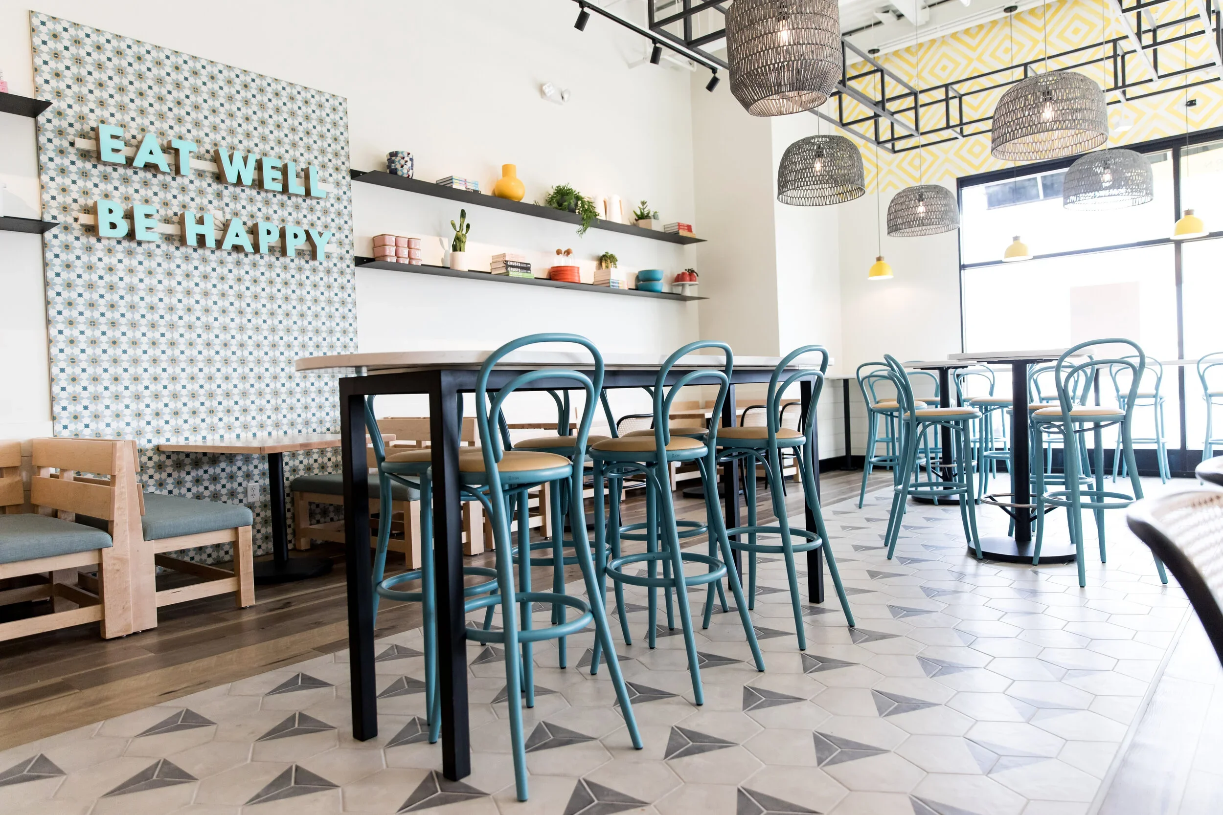 Restaurant communal dining with tall wood table, blue bar stools, gray woven pendants, patterned floor tile, and greenery. Photography by Nathan Warmes