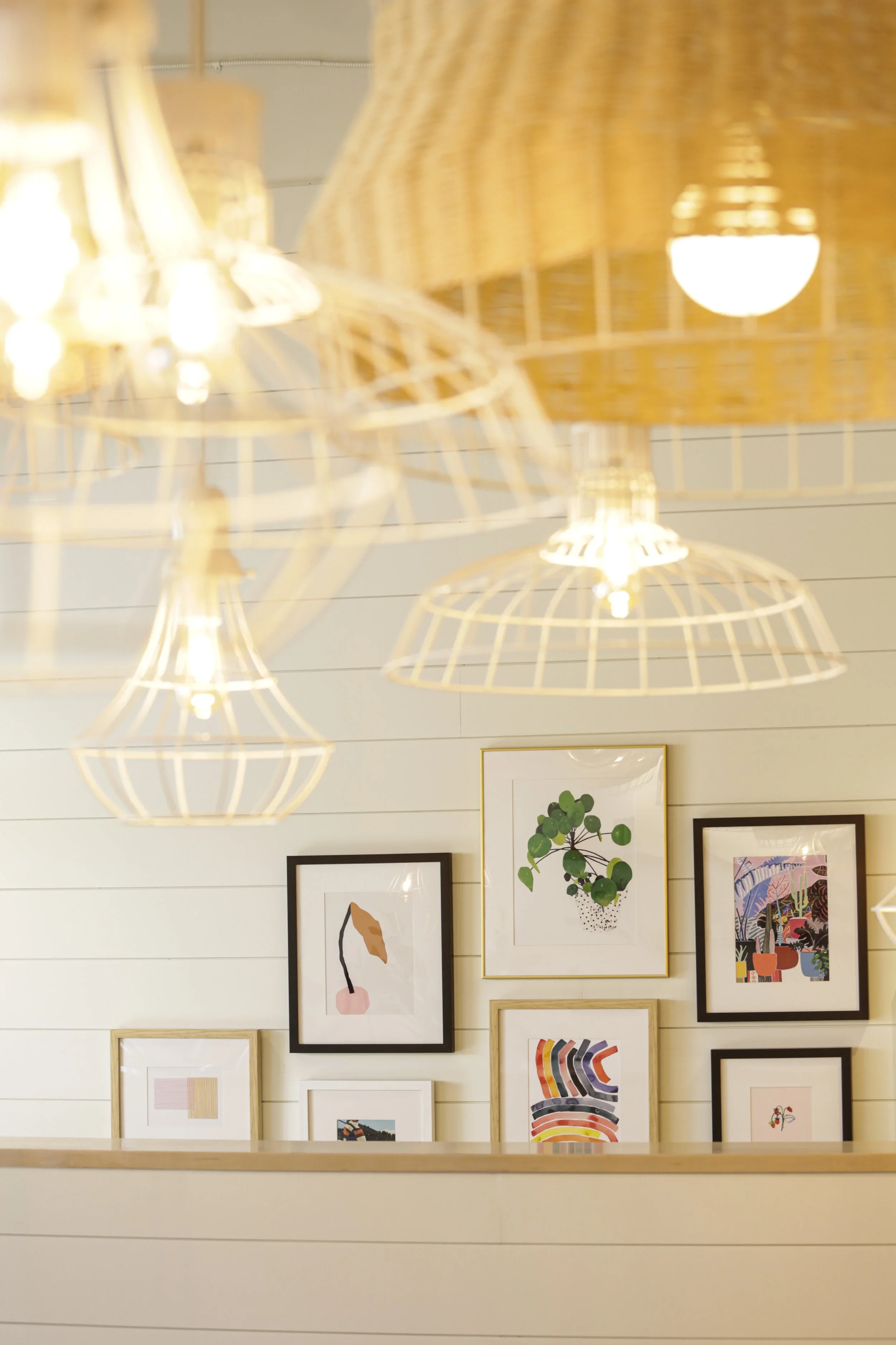 Restaurant lighting detail with white wire-frame and pleated pendant shades above framed artwork on shiplap walls. Photography by Nathan Warmes