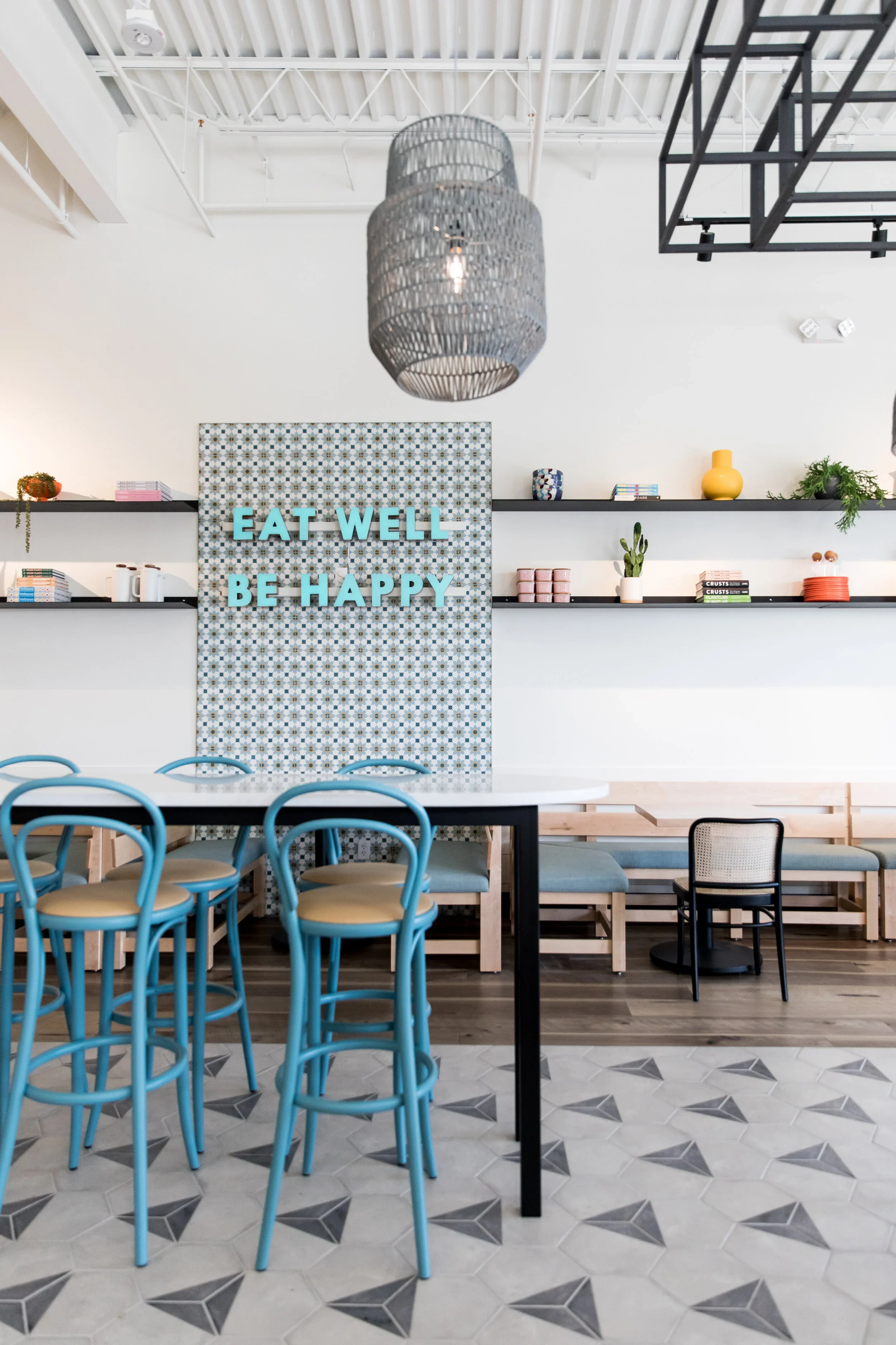 Restaurant banquette with central blue patterned tile, pale wood seating, cane-back chairs, and shelves of bowls and plants. Photography by Nathan Warmes