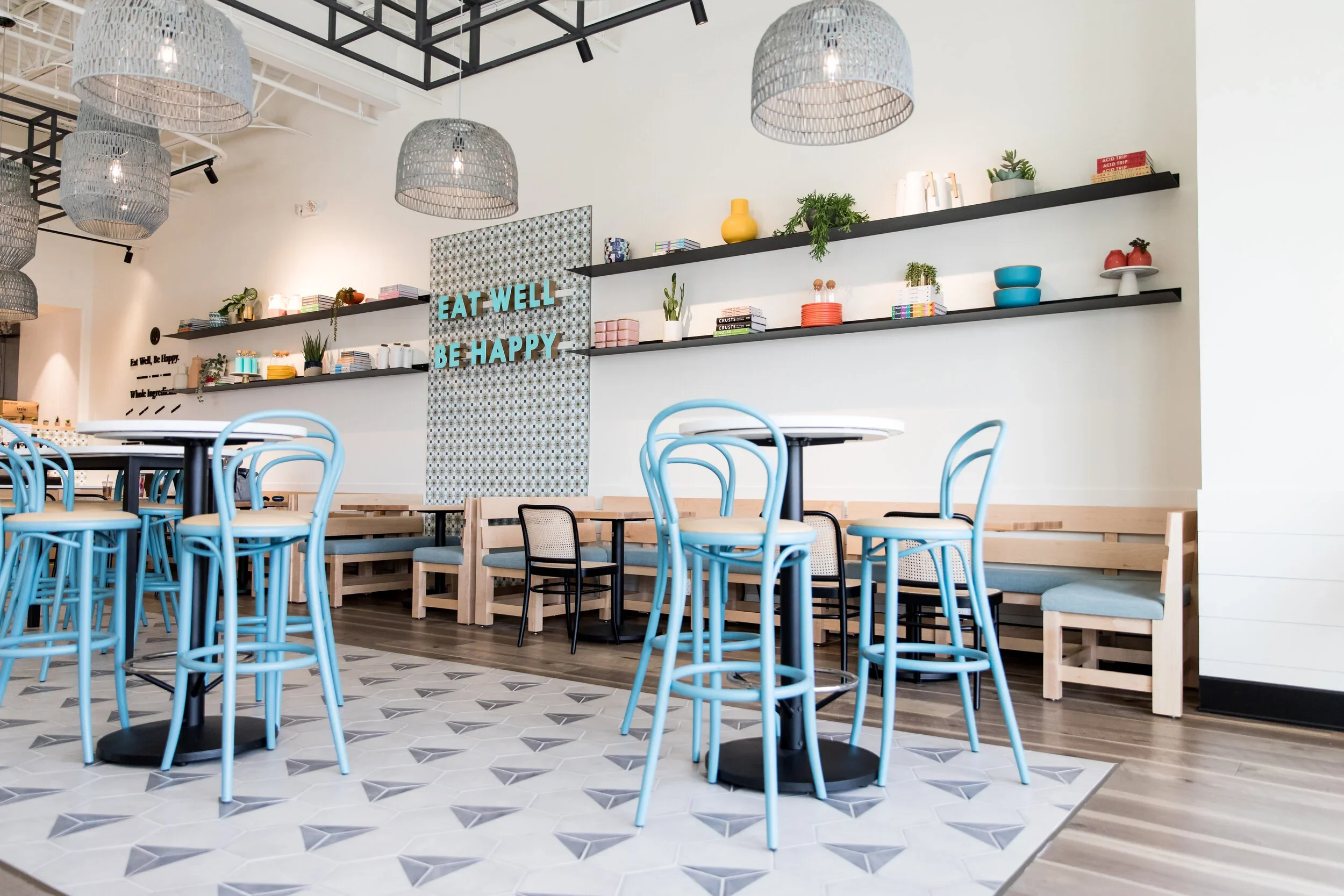 Restaurant dining room with blue chairs, wood banquette, graphic floor tile, globe pendants, and shelves of ceramics and plants. Photography by Nathan Warmes