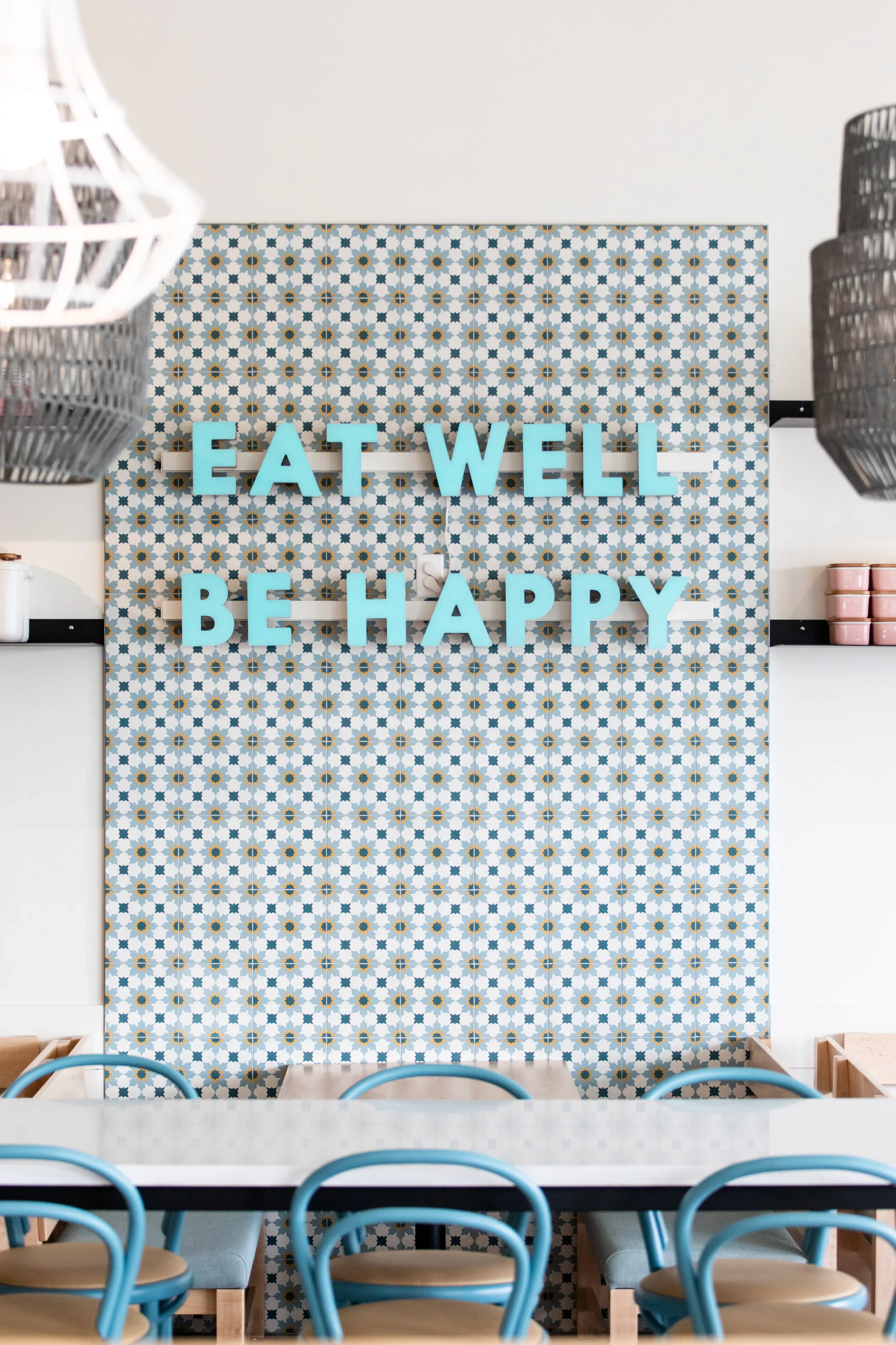 Restaurant feature wall with blue geometric tile, turquoise 'Eat Well Be Happy' lettering, wood banquette, and blue chairs. Photography by Nathan Warmes