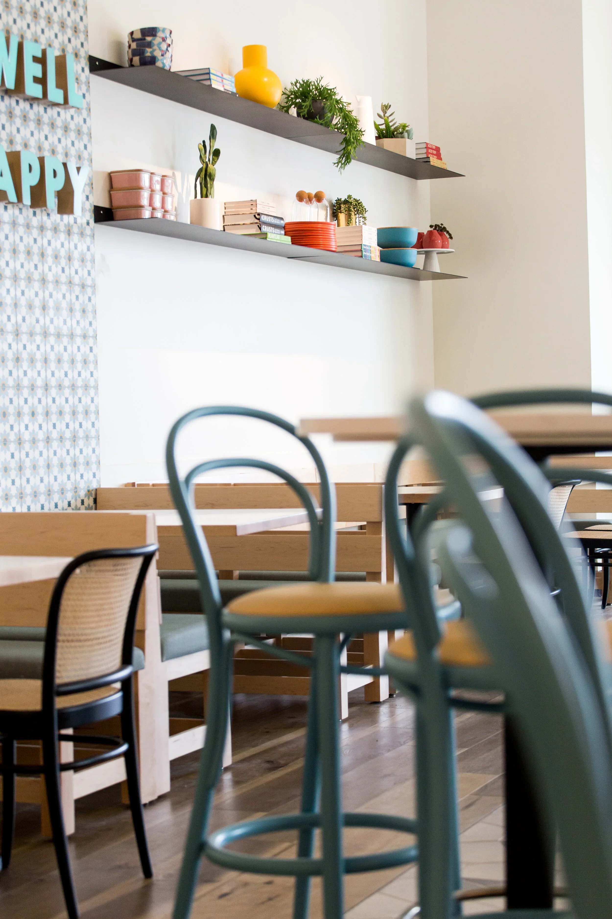 Restaurant seating detail with blue bentwood chairs, wood banquette, white shiplap wall, and shelf of colorful dishes and plants. Photography by Nathan Warmes