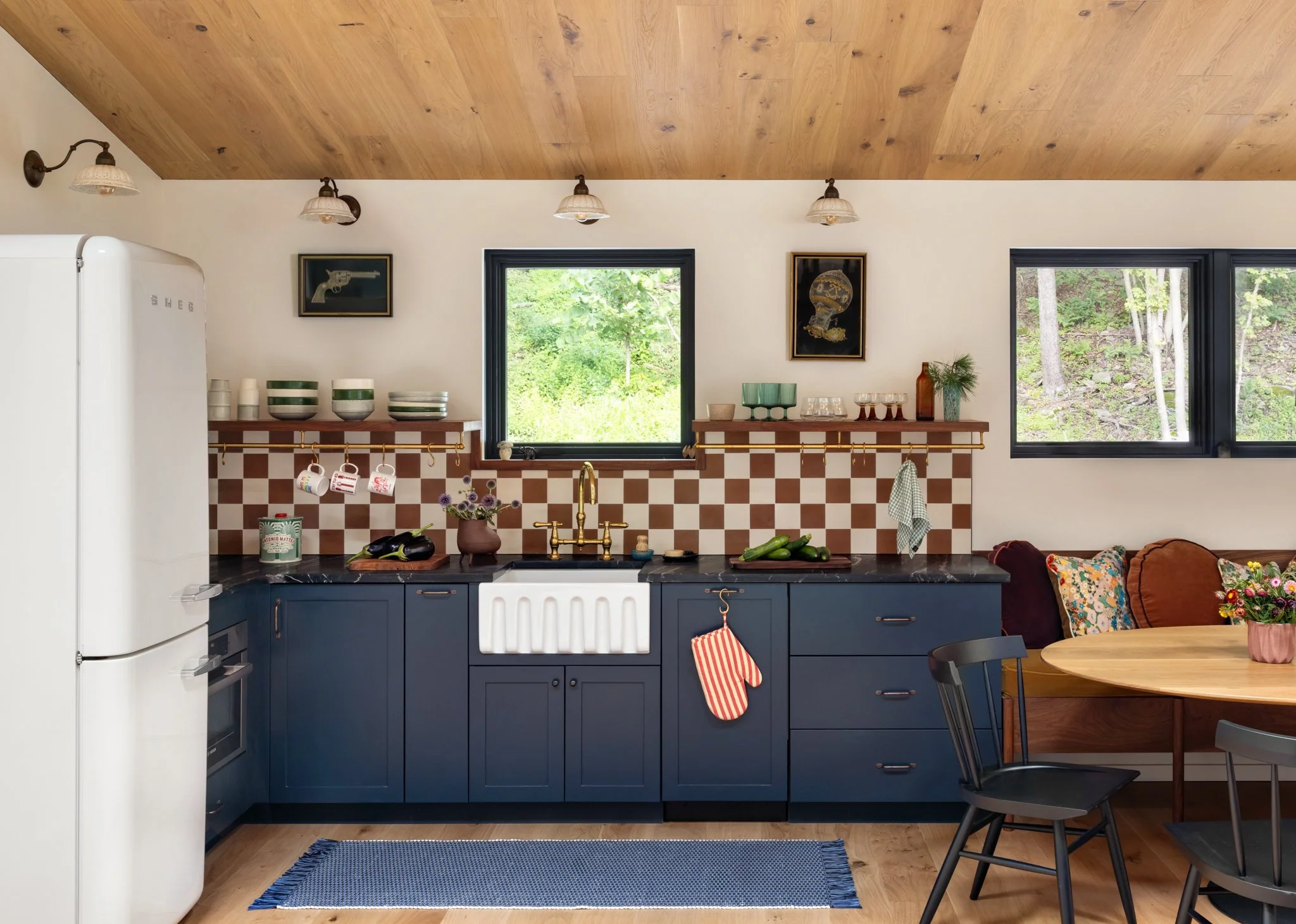 Guest-house kitchen with navy cabinets, burgundy-and-cream checkerboard tile, white farmhouse sink, and a timber ceiling.