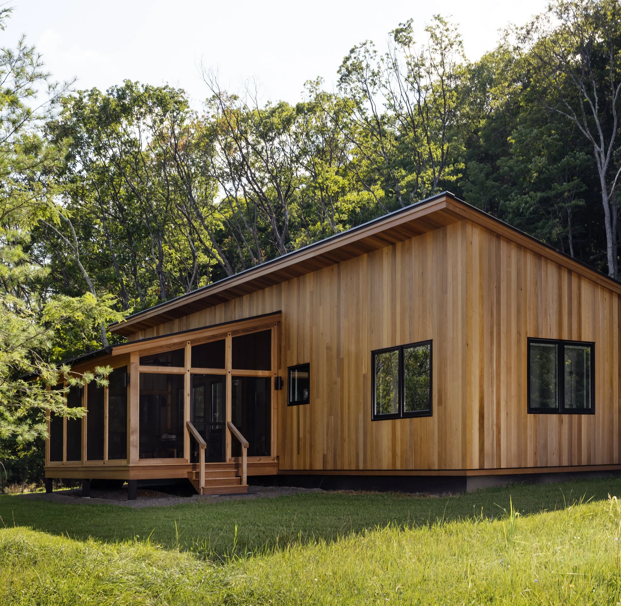 Cedar-clad guest house with a pitched roof, screened porch, dark-framed windows, and surrounding woodland.