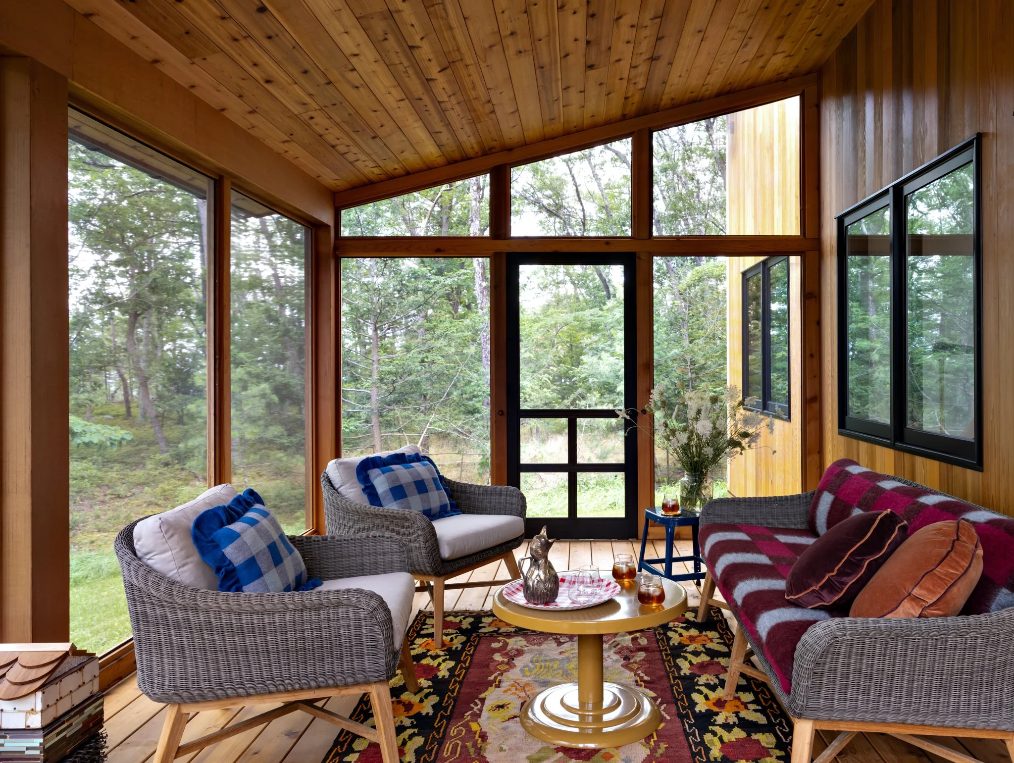 Screened porch with a timber ceiling, patterned seating, blue checked cushions, a yellow side table, and woodland views.