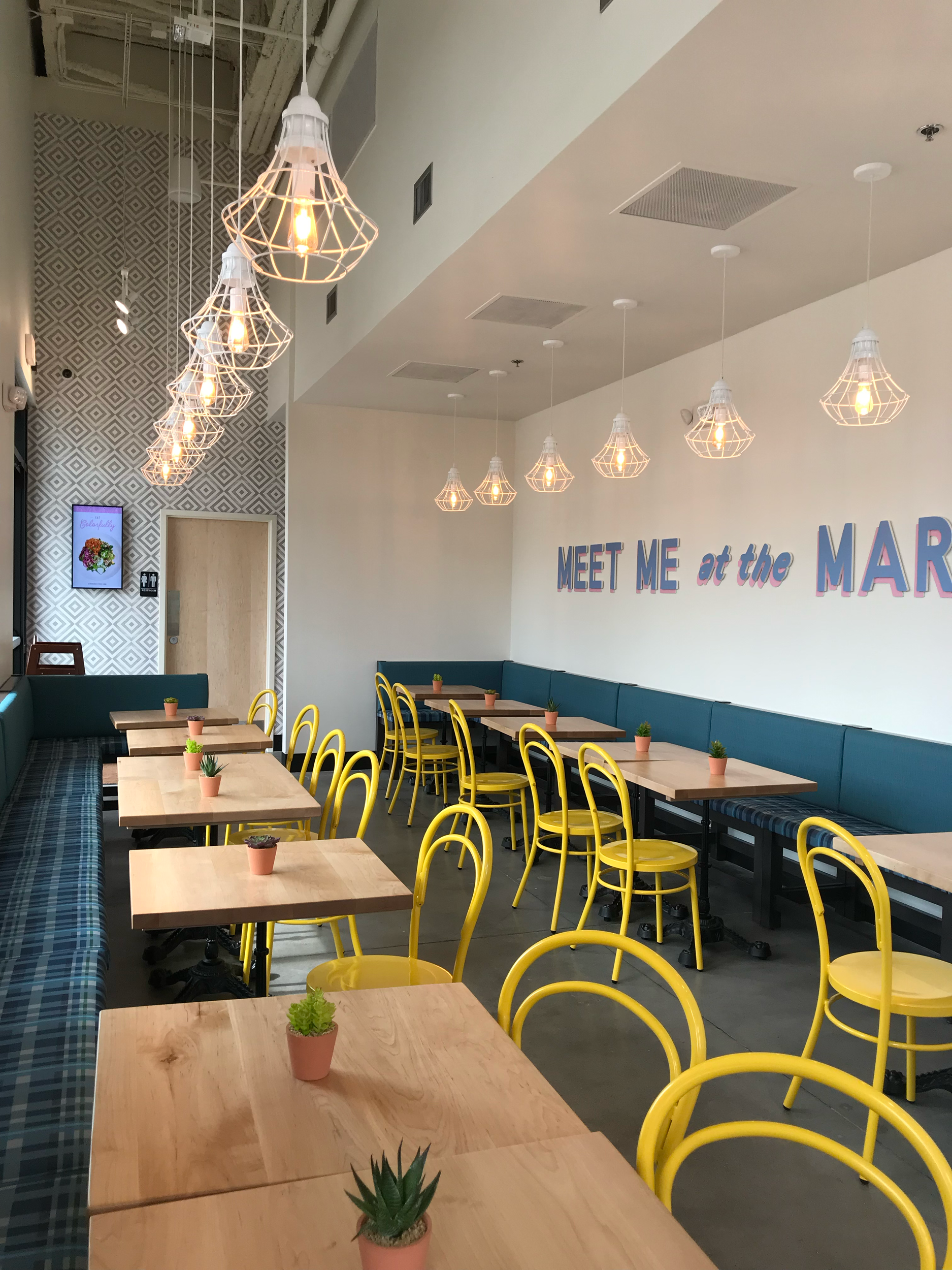 Restaurant dining area with yellow chairs, wood tables, blue banquettes, and pendant lights.
