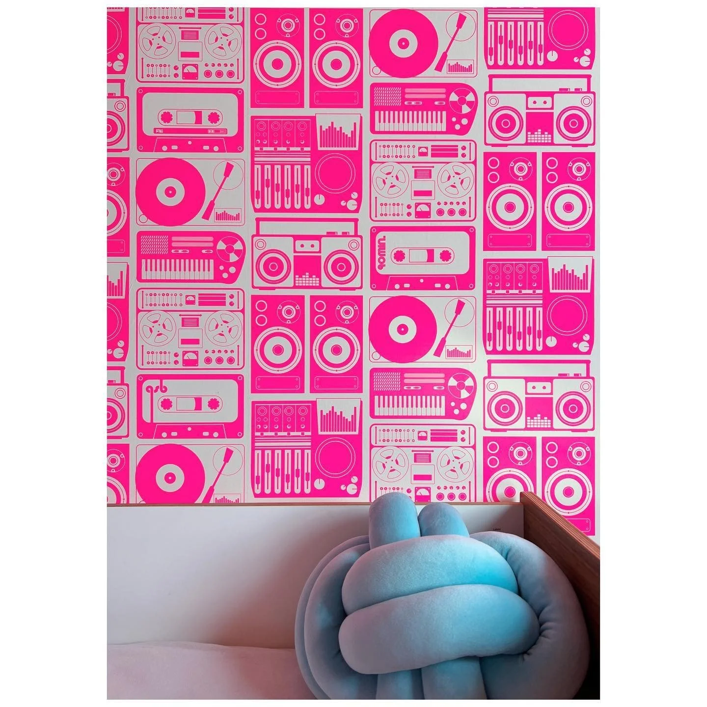Bright pink illustrated wallpaper above a pale upholstered bed and blue knot cushion