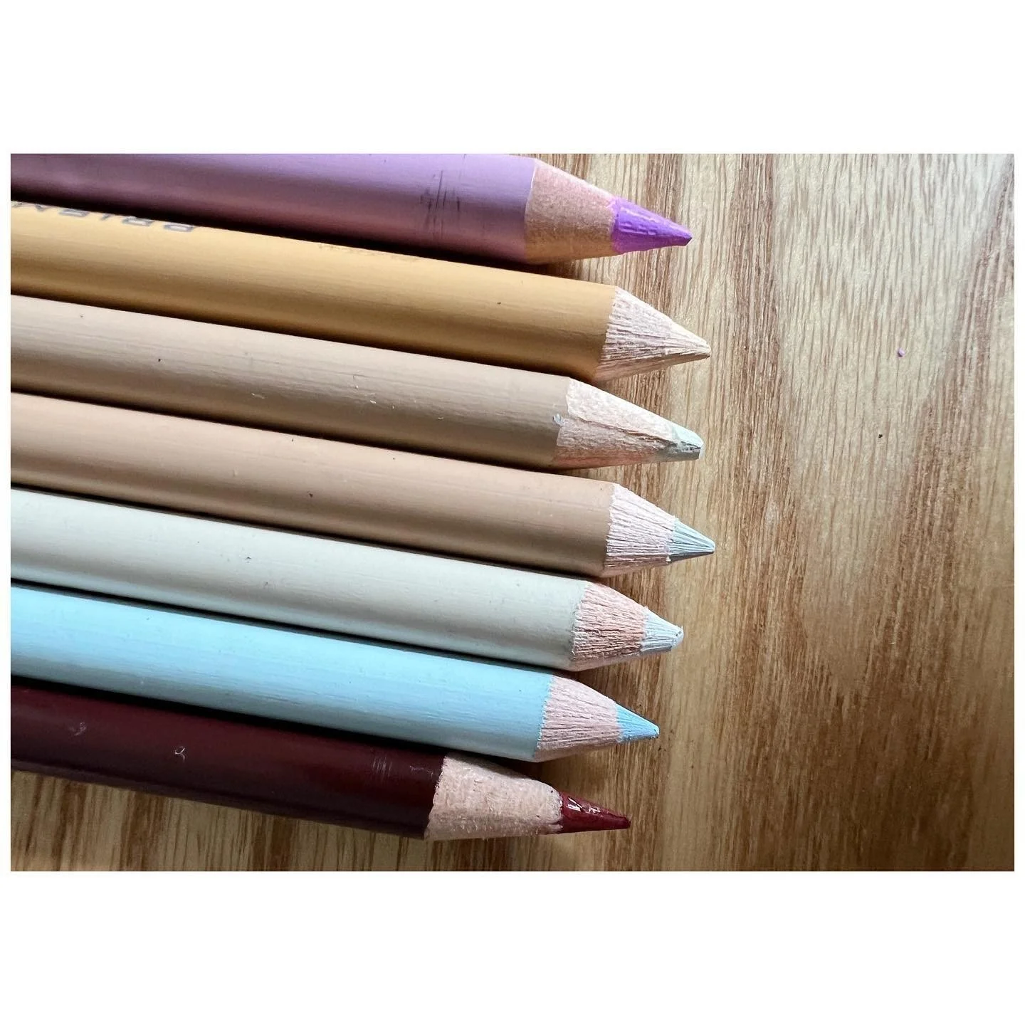 Colored pencils in soft pink green blue and brown arranged over a pale wood surface