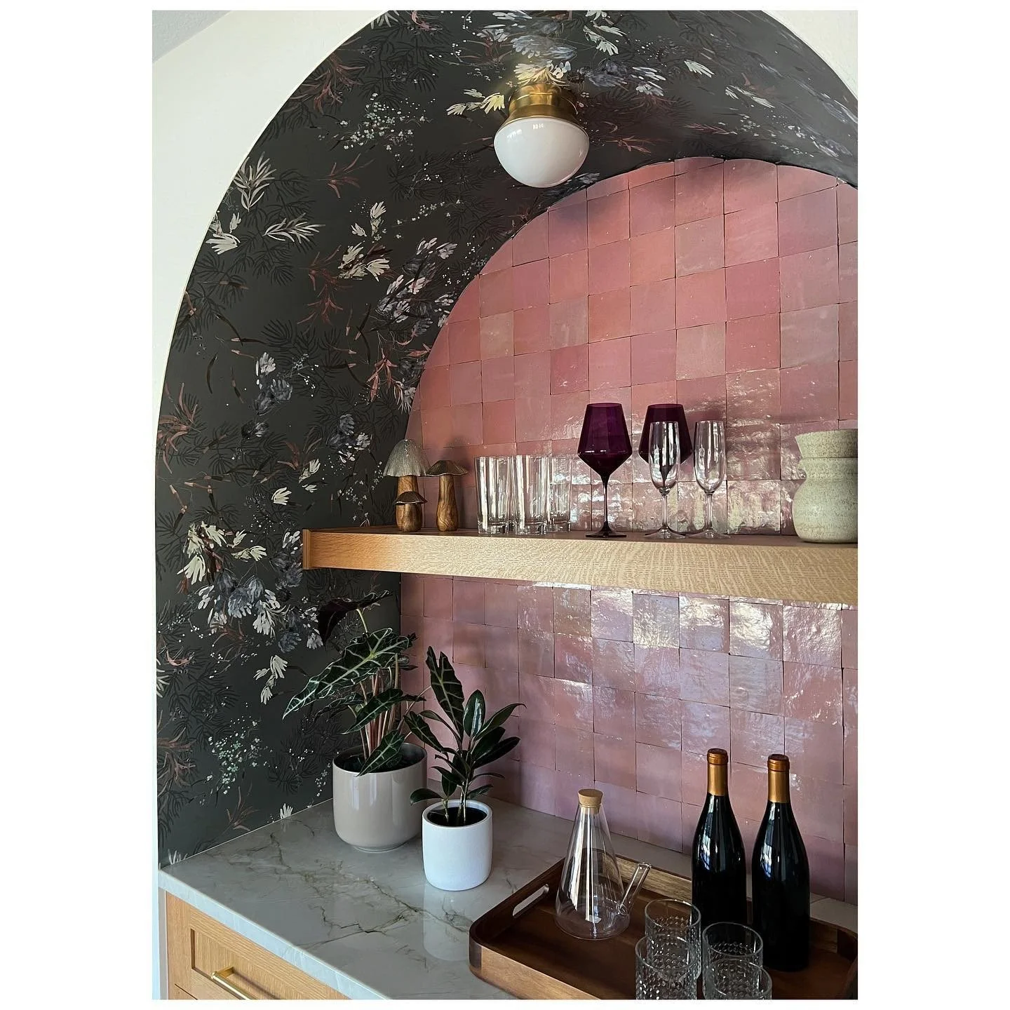 Glassware arranged inside a curved niche with dark floral wallpaper and a pink backing