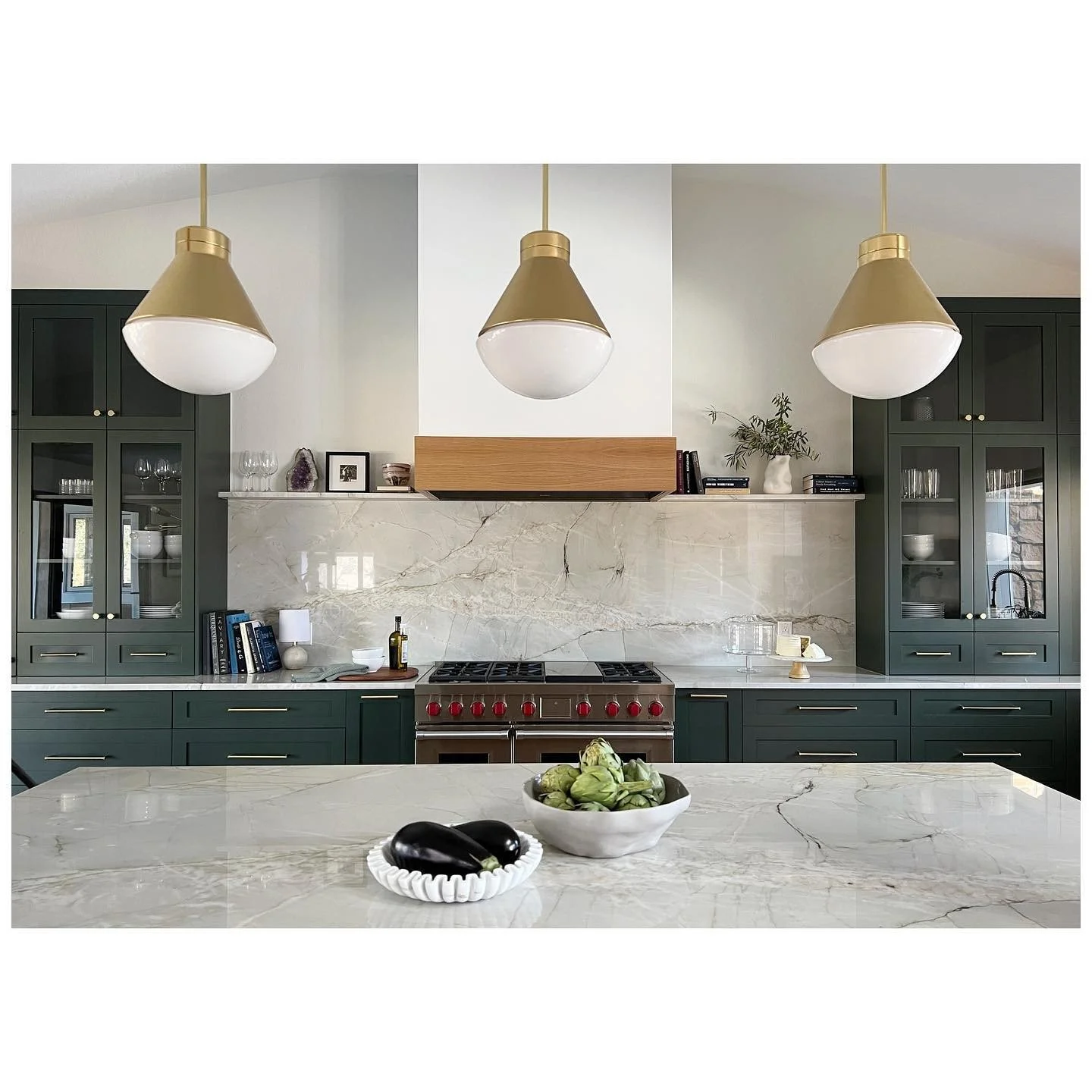 Kitchen island and three brass pendant lights against pale cabinetry and a dark range hood