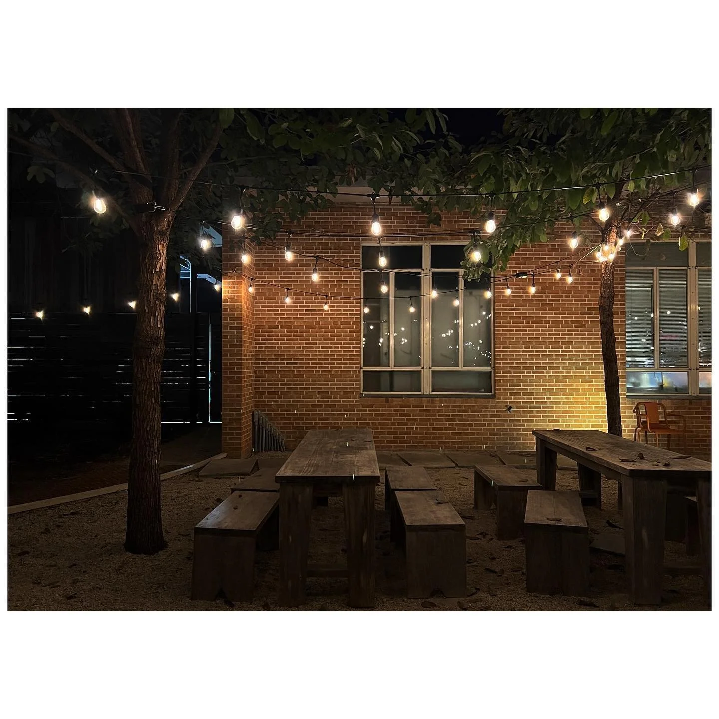 Outdoor dining tables beneath warm string lights beside a brick building at night