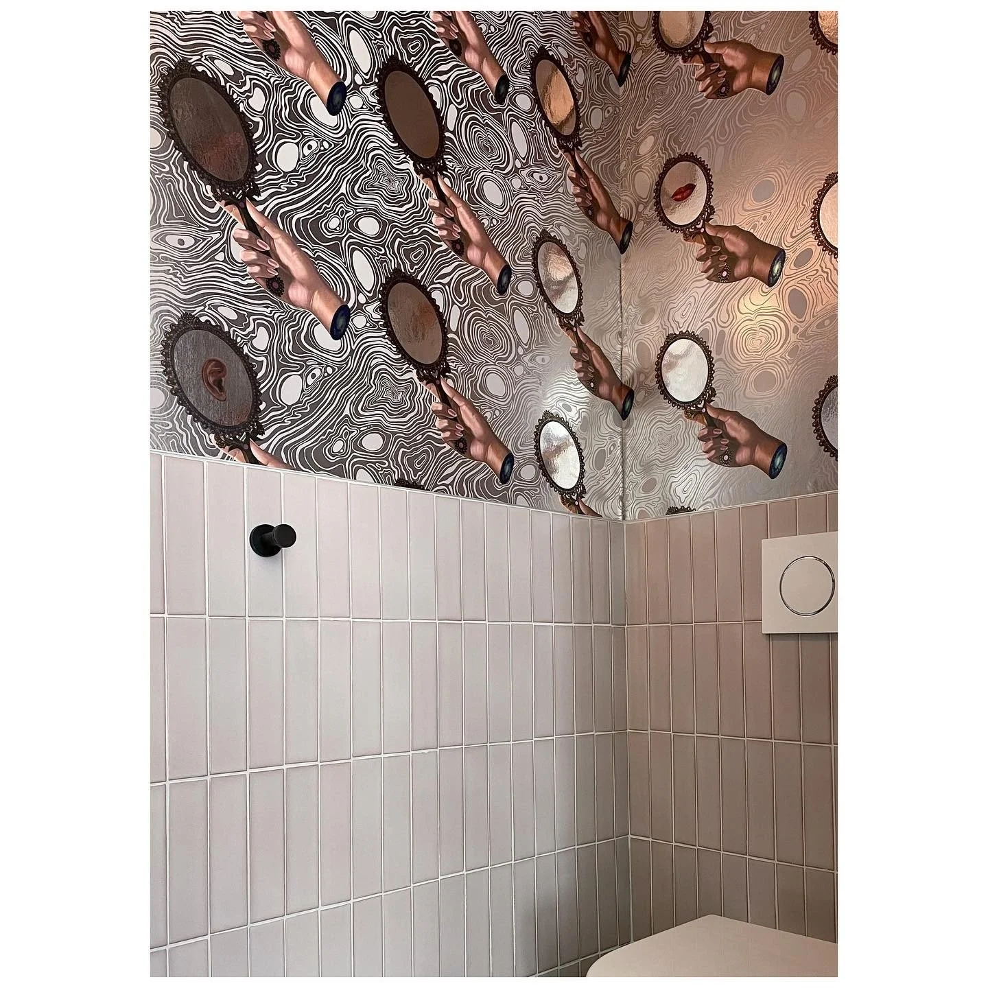 Patterned wallpaper with illustrated frames above pale rectangular bathroom tiles and dark fixtures