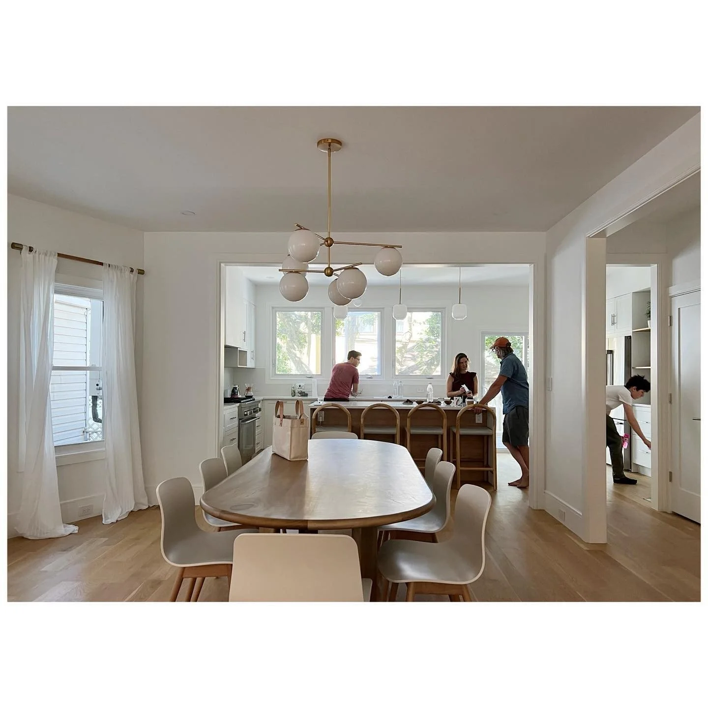 Light-filled dining room with a timber table, pale chairs and a brass globe pendant opening into a kitchen during a project visit