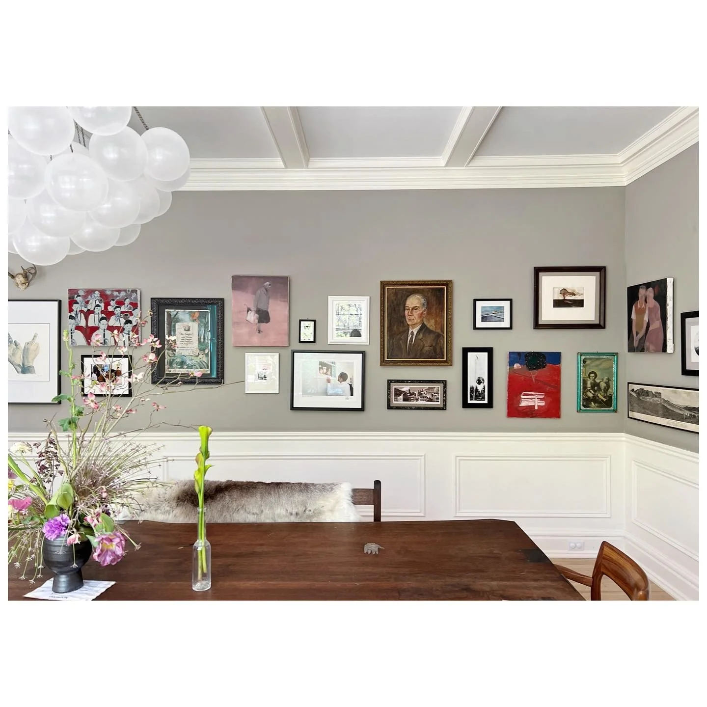 Collected framed artworks across a gray dining room wall above white paneling and a timber dining table