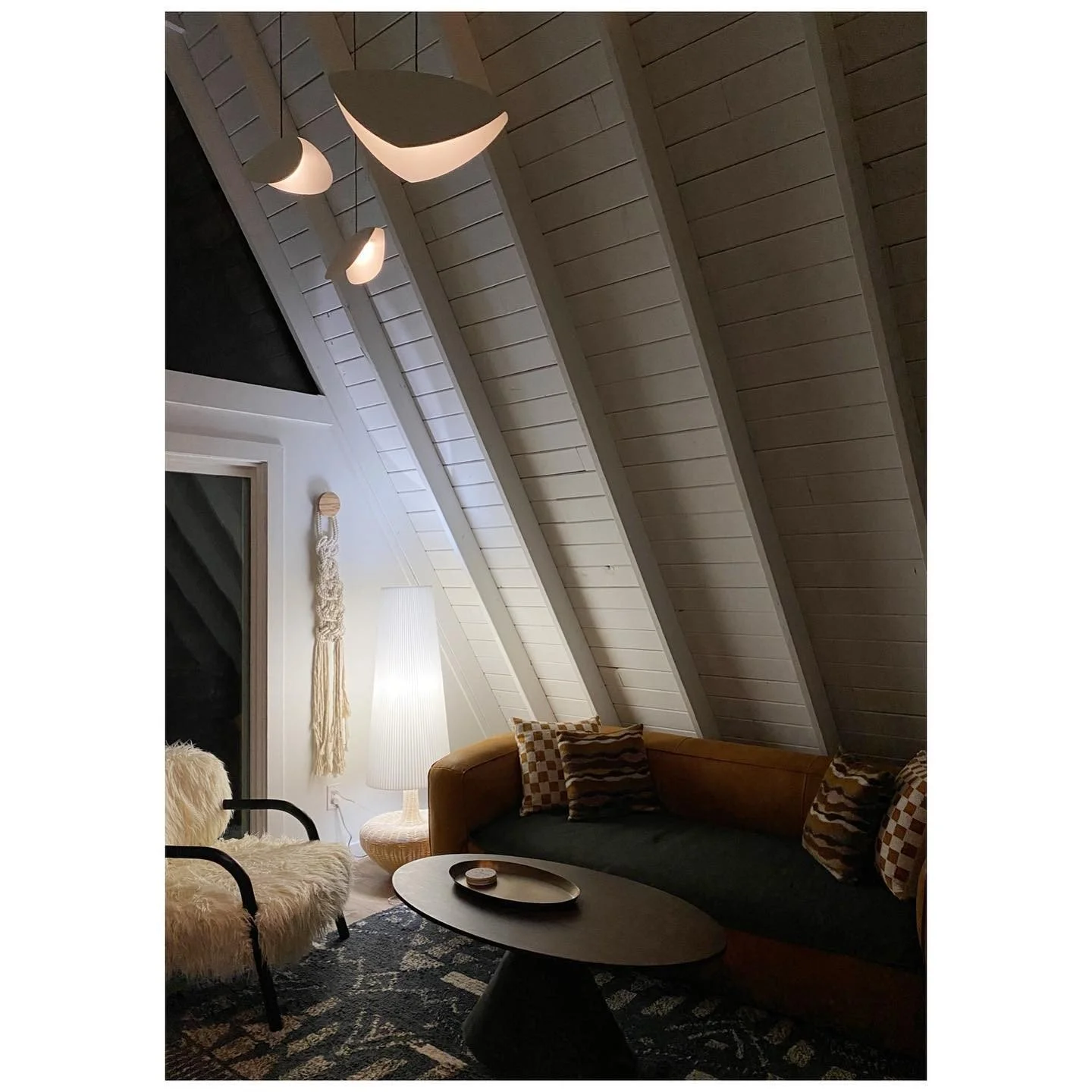 Moody cabin living room with angled white roof beams, hanging sculptural lights, a mustard sofa and woven wall art