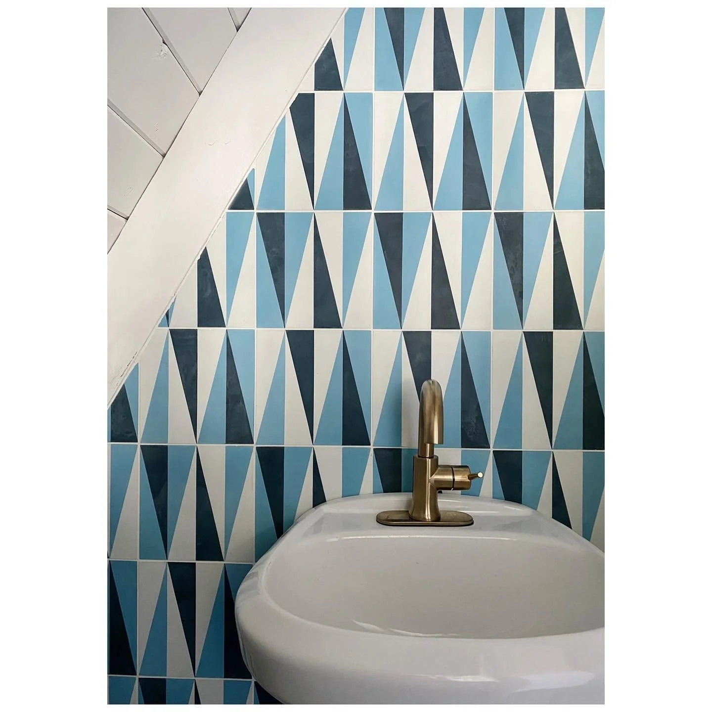 Round white bathroom basin with brass tap in front of blue-and-white geometric wallpaper beneath an angled roof