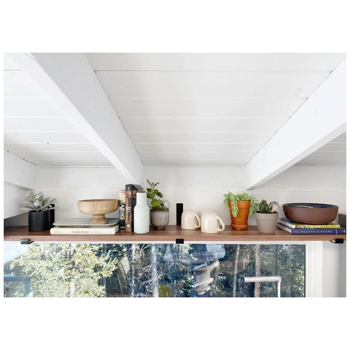 Timber shelf styled with ceramic bowls, mugs, utensils and plants above a wide cabin kitchen window