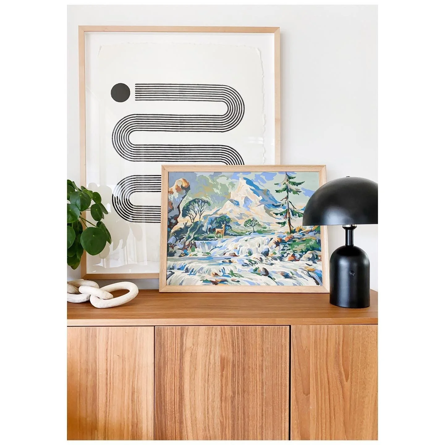 Framed landscape painting and graphic artwork styled above a timber cabinet with a black lamp