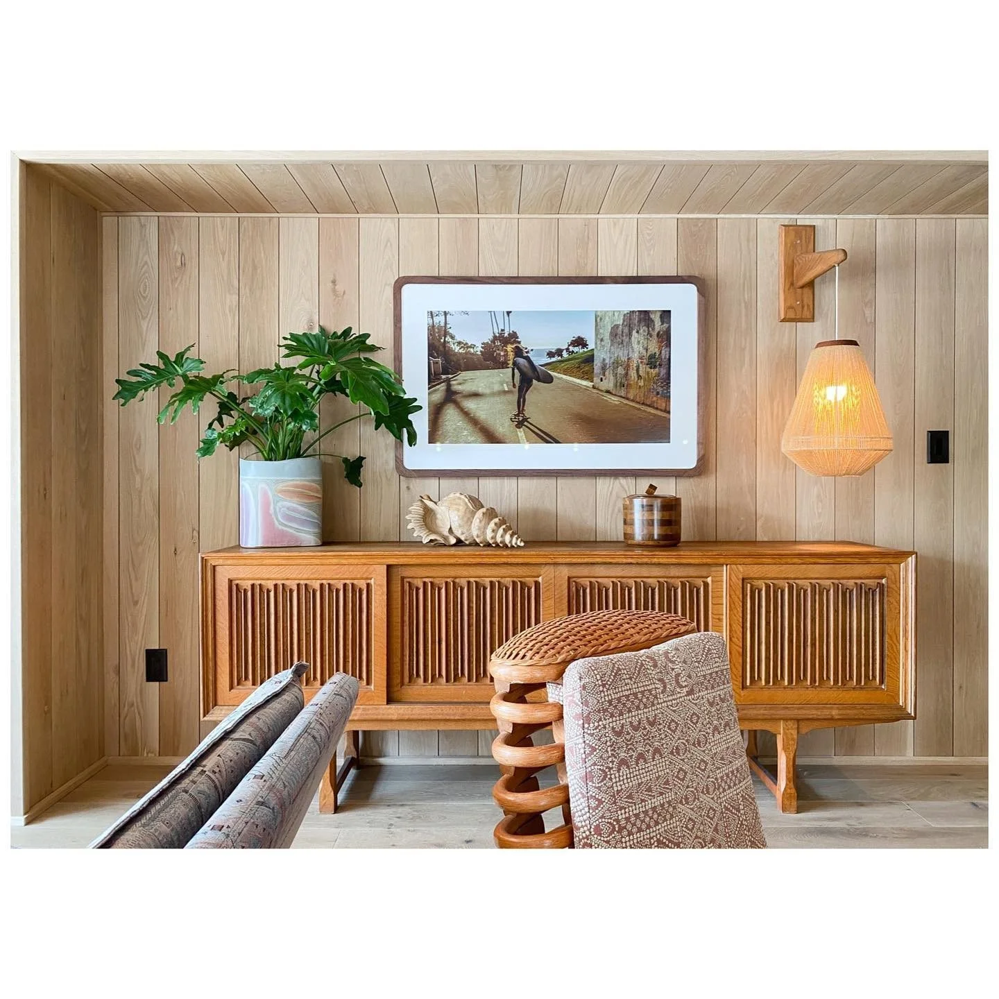 Timber-paneled sitting room with a slatted credenza, framed artwork, leafy plants and a woven wall light