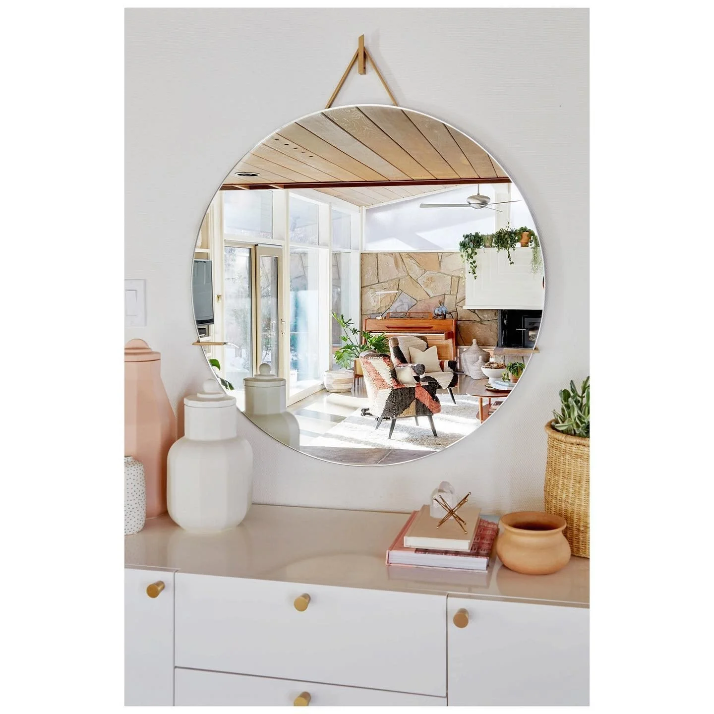 White entry cabinet with a round hanging mirror, sculptural white vases, stacked books and a reflection of a bright dining room