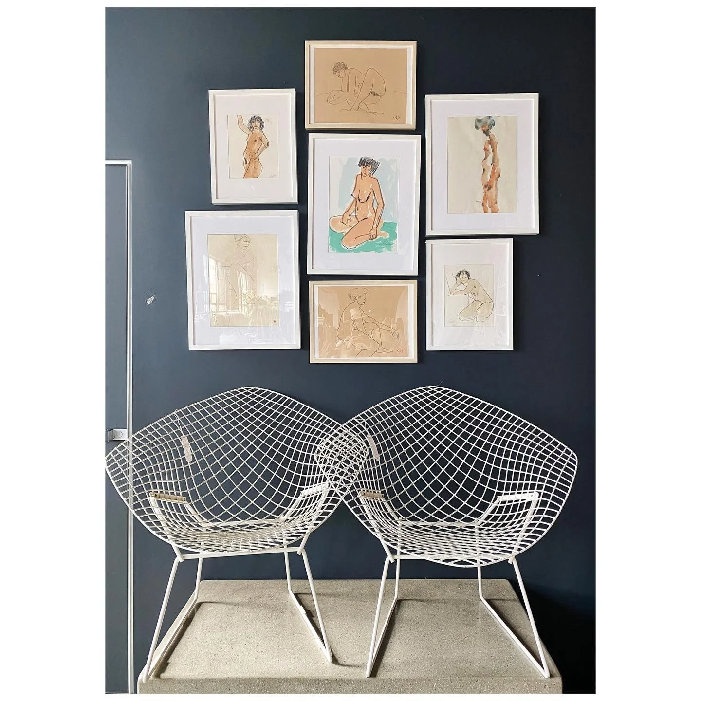 Framed figure drawings arranged above two white wire lounge chairs against a dark blue wall